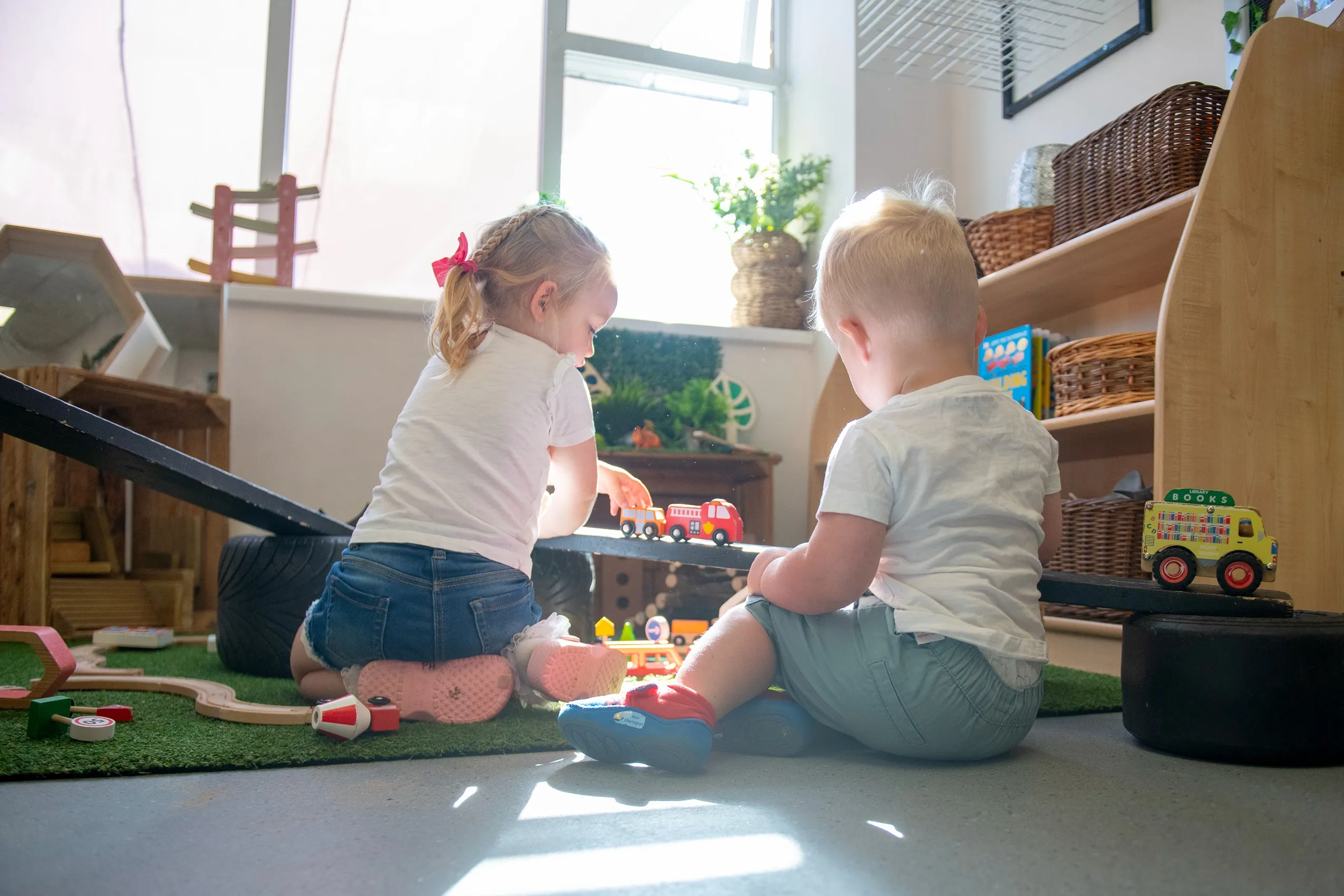 Two young children, a girl with a pink bow and a boy, playing with a toy train and a yellow toy bus.