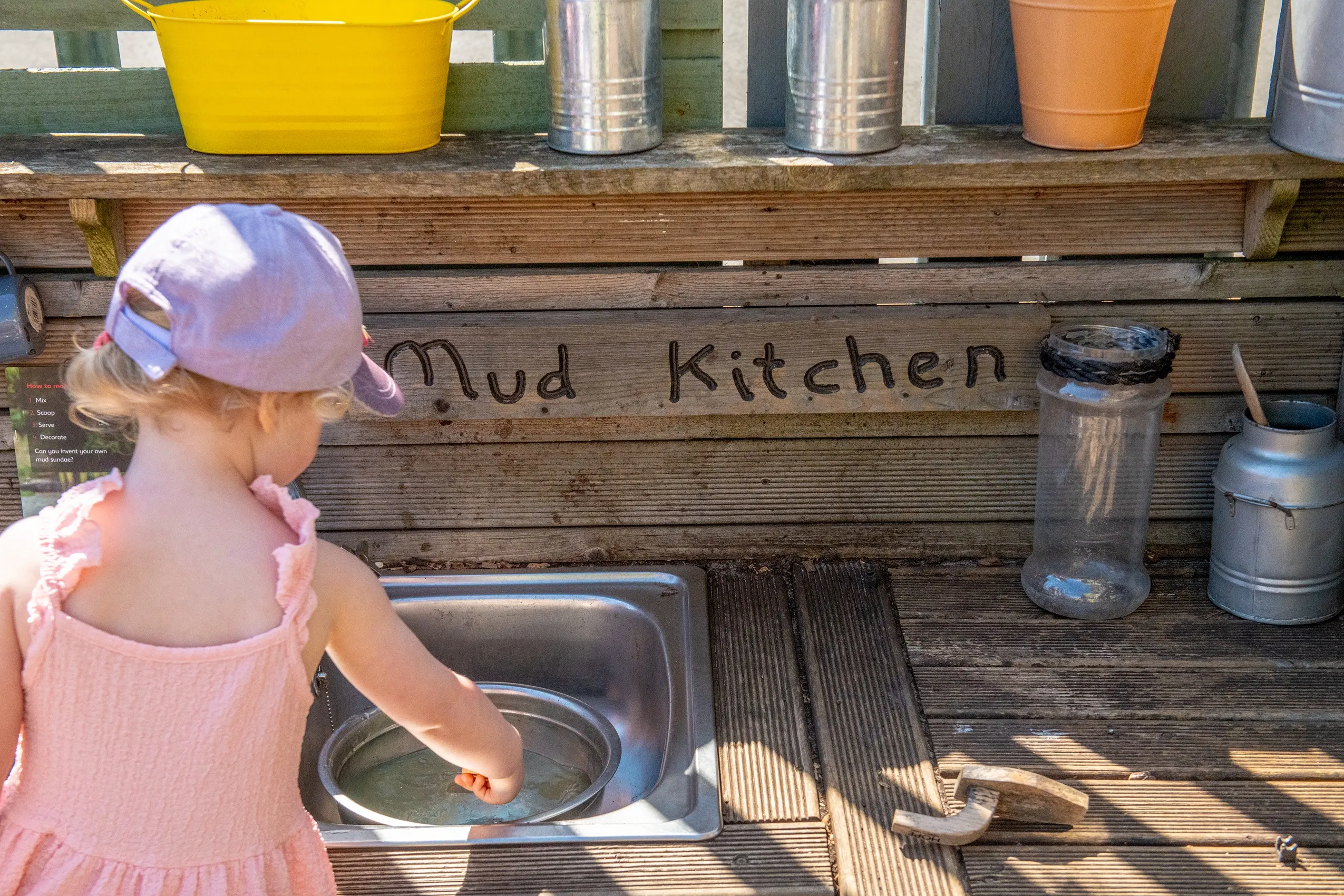 A wooden mud kitchen with a metal sink, surrounded by containers and utensils, with a child playing at it.