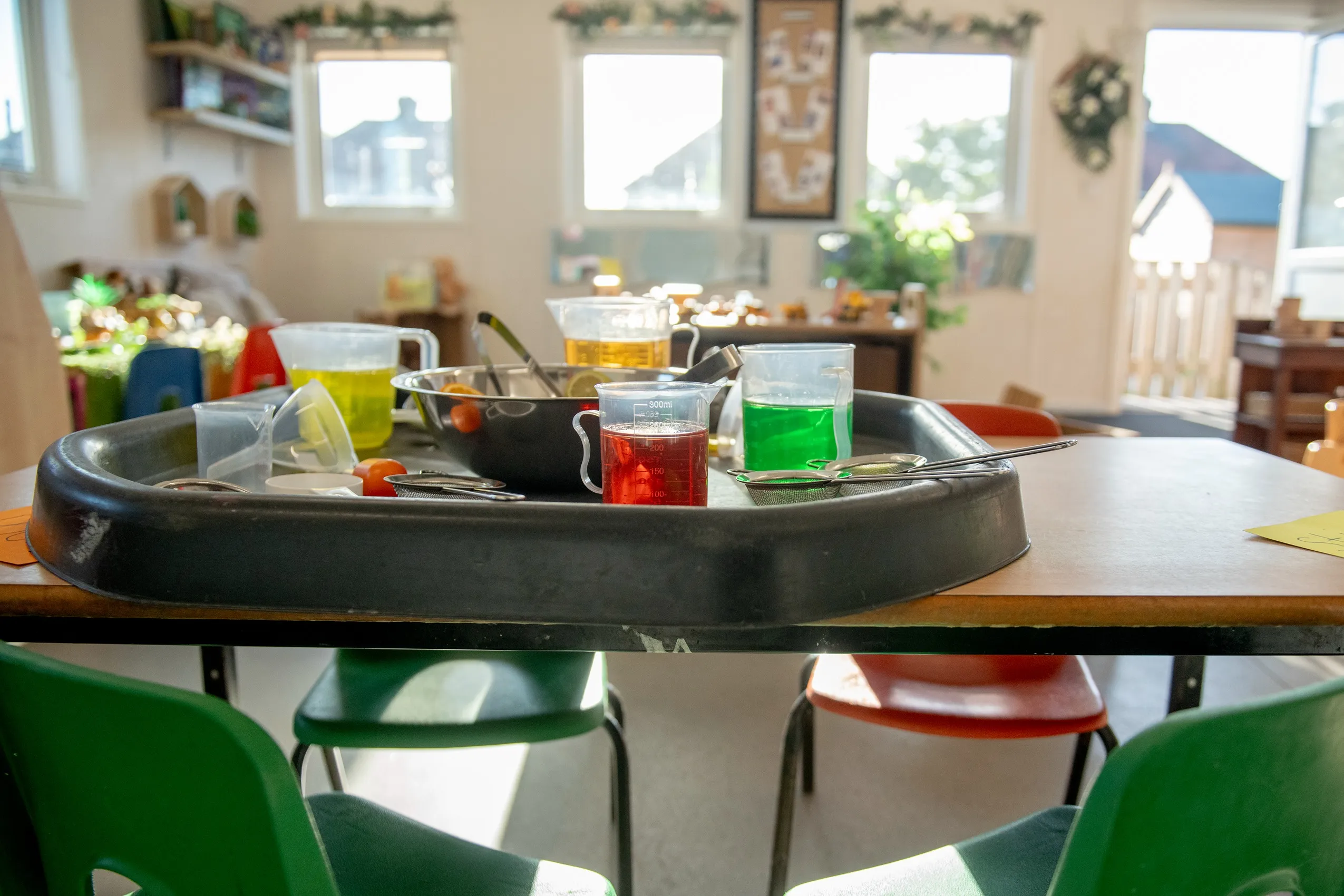 A black plastic tray filled with colorful liquids in pitchers and measuring cups, with a metal bowl and utensils, set up for an experiment.