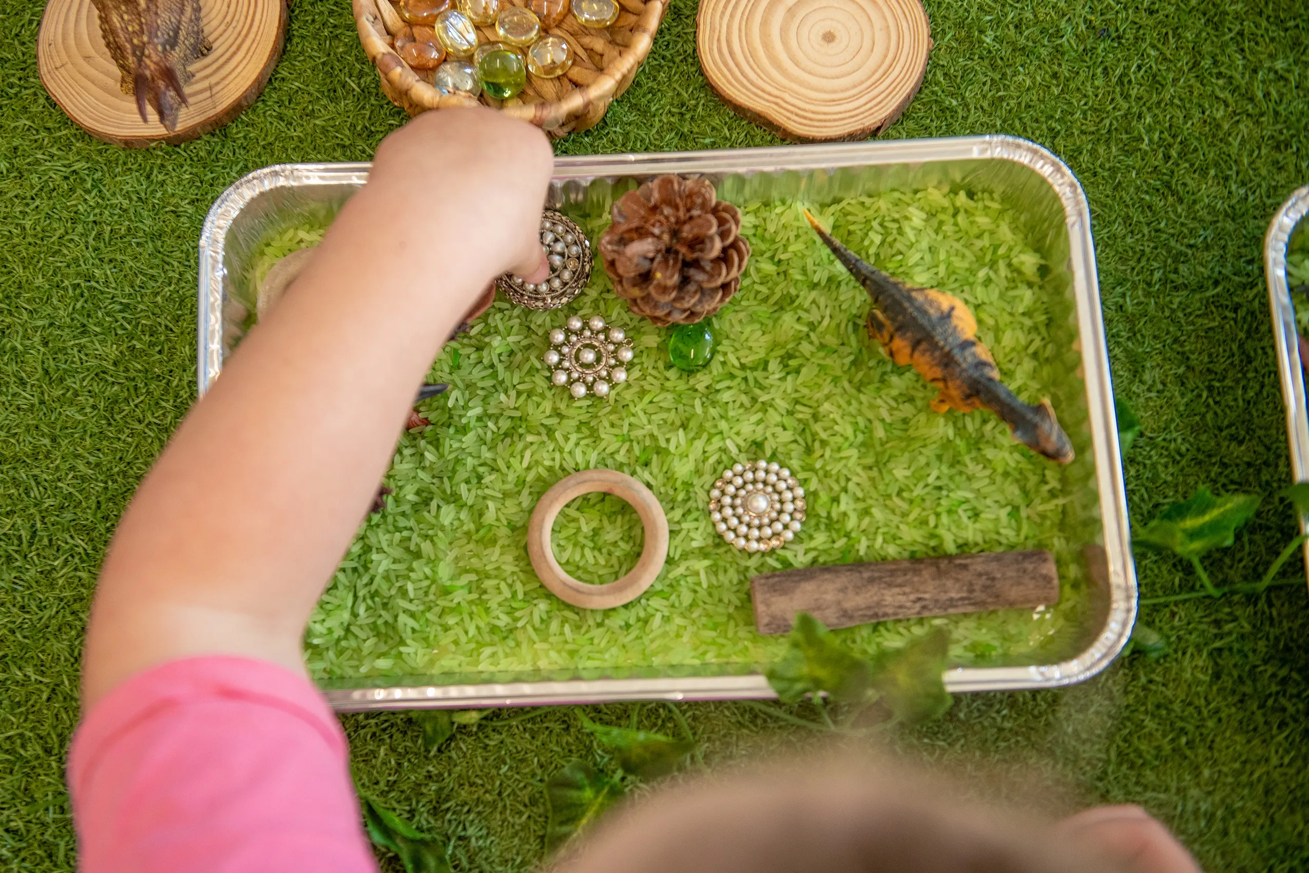 A child's hand interacting with a sensory bin filled with green rice, pine cones, and toy dinosaurs.