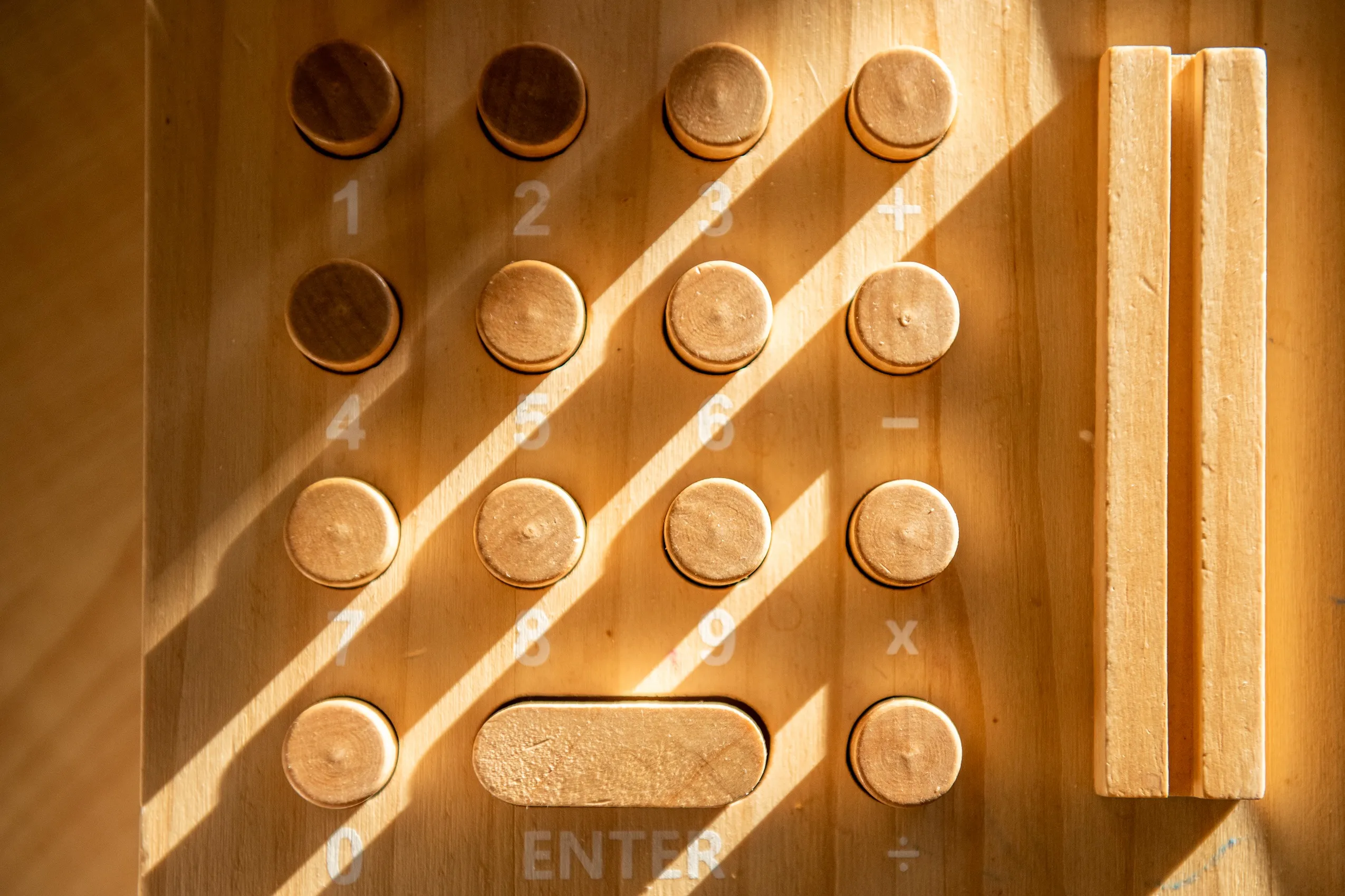 A wooden calculator with a grid of numbered buttons and mathematical symbols, lit by sunlight.