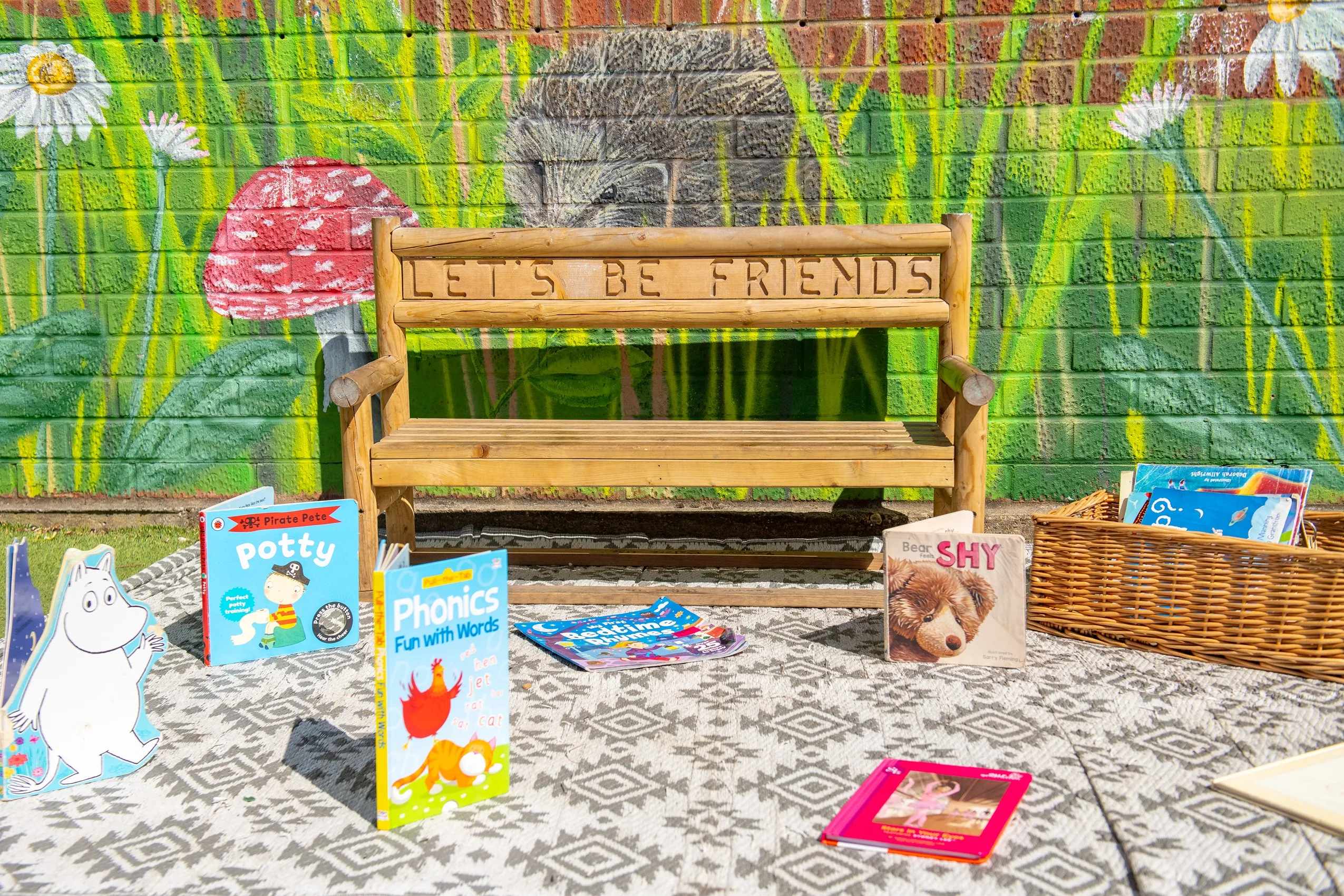 A wooden bench with 'LET'S BE FRIENDS' carved into it, in front of a colorful brick wall with children's books.