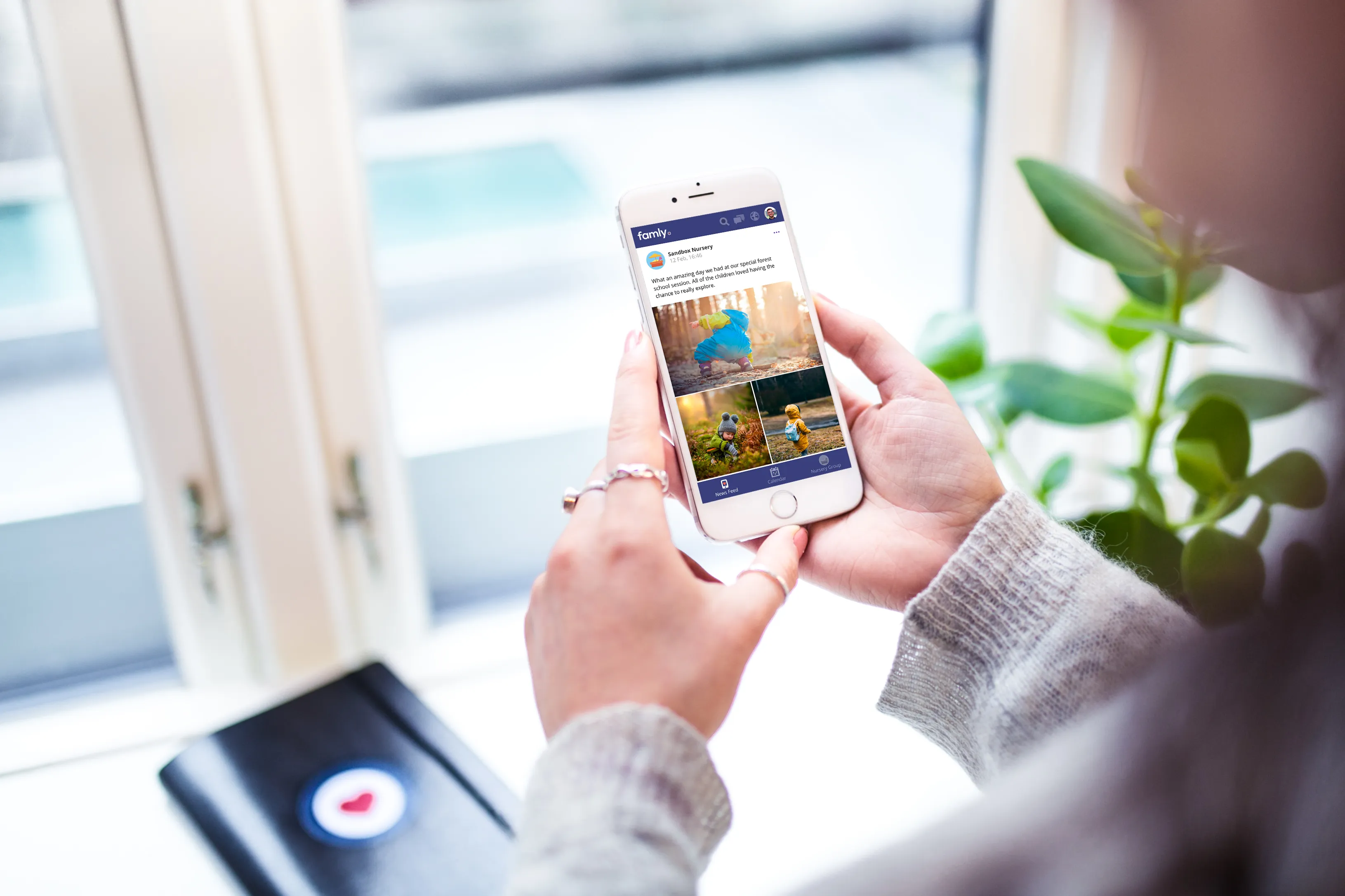 A person holding a white smartphone displaying a 'famly' social media feed with photos of children playing outdoors.