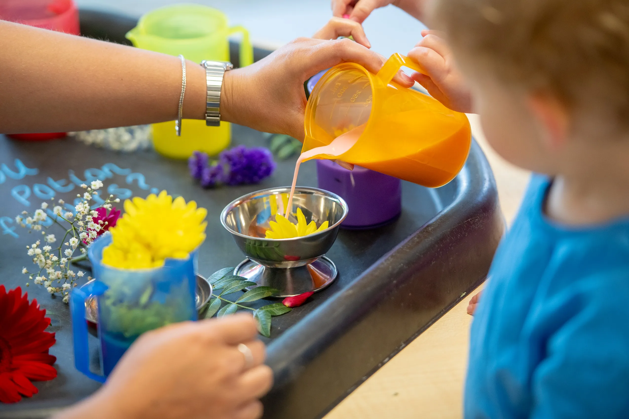 A yellow measuring cup pouring pink liquid into a bowl with a yellow flower, surrounded by colorful items.