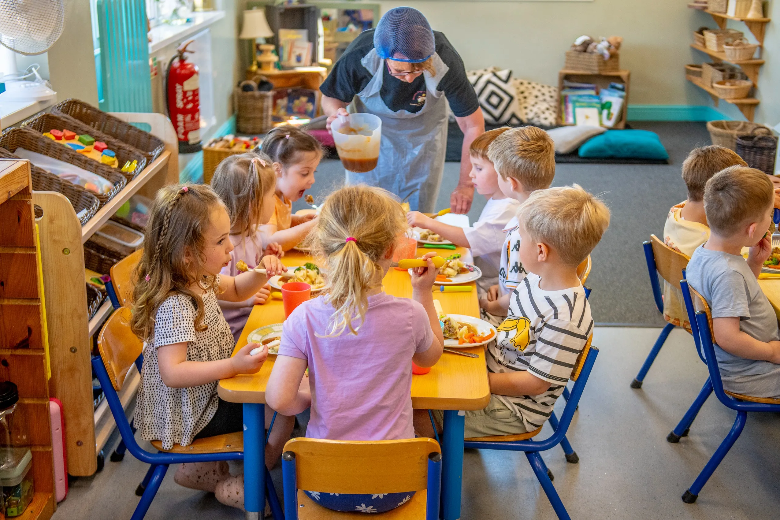 A group of young children sitting around a yellow table, eating lunch, with a woman serving them.