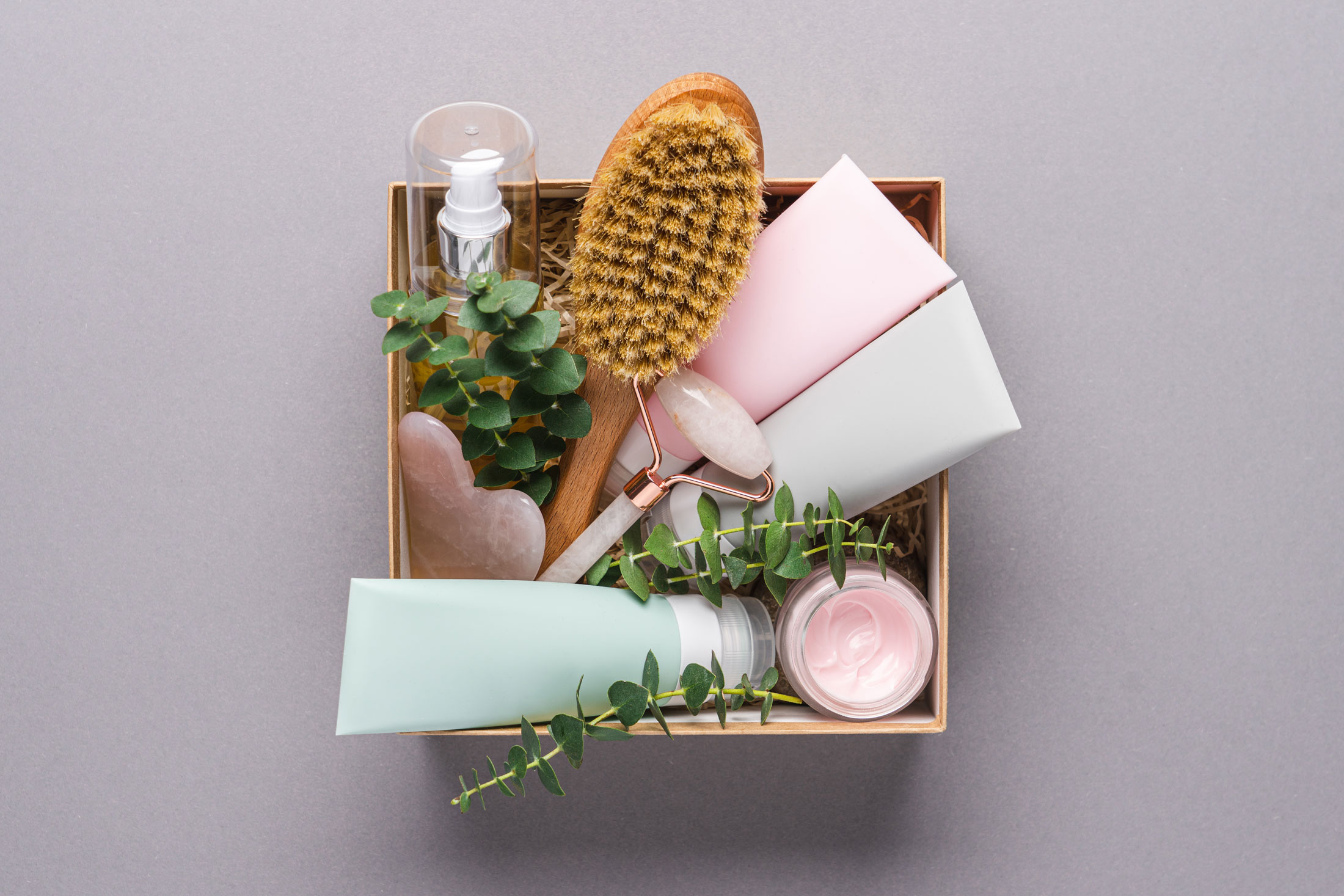Box with skincare items: tubes, brush, jade roller, and small plants arranged neatly.