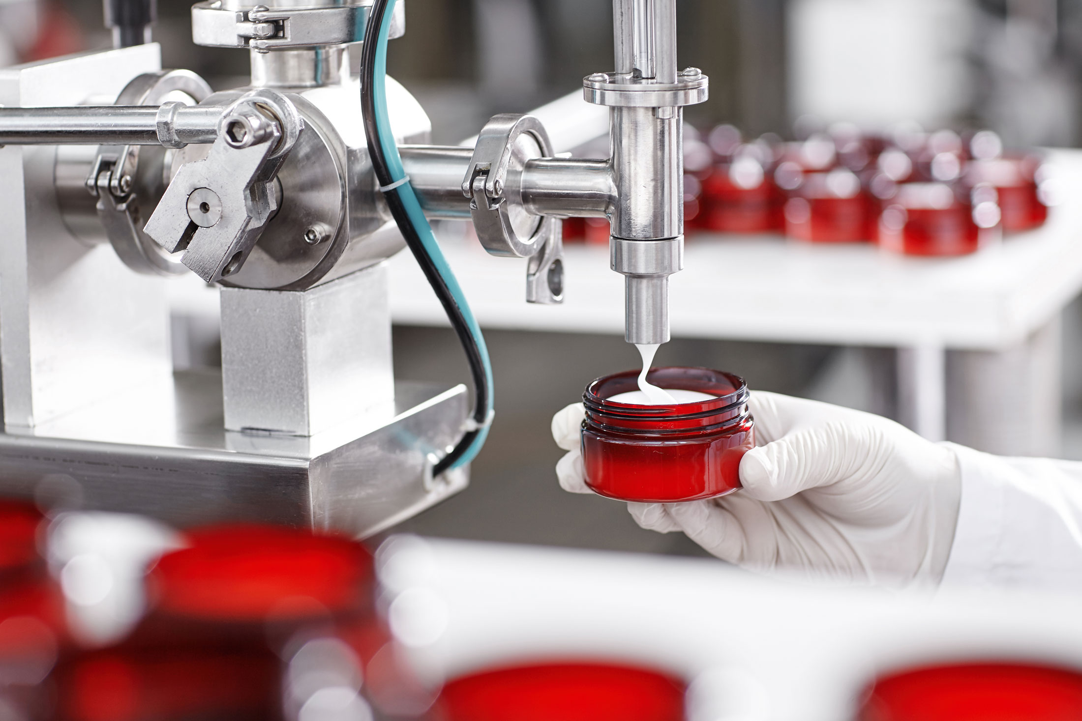 A gloved hand holds a red jar being filled with cream by a machine.