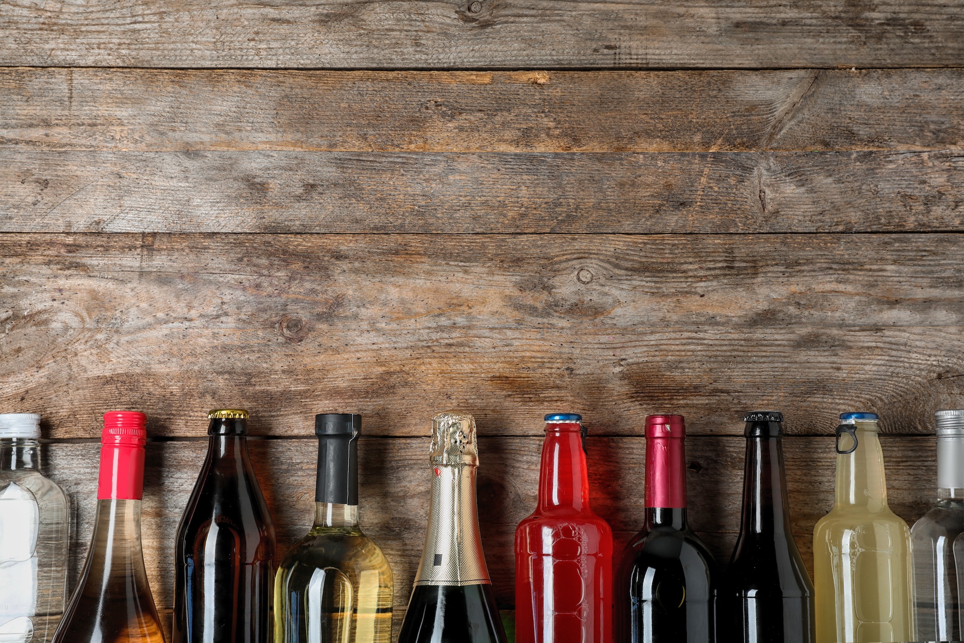 A row of various bottles, including wine, liquor, and beer, in front of a wooden background.