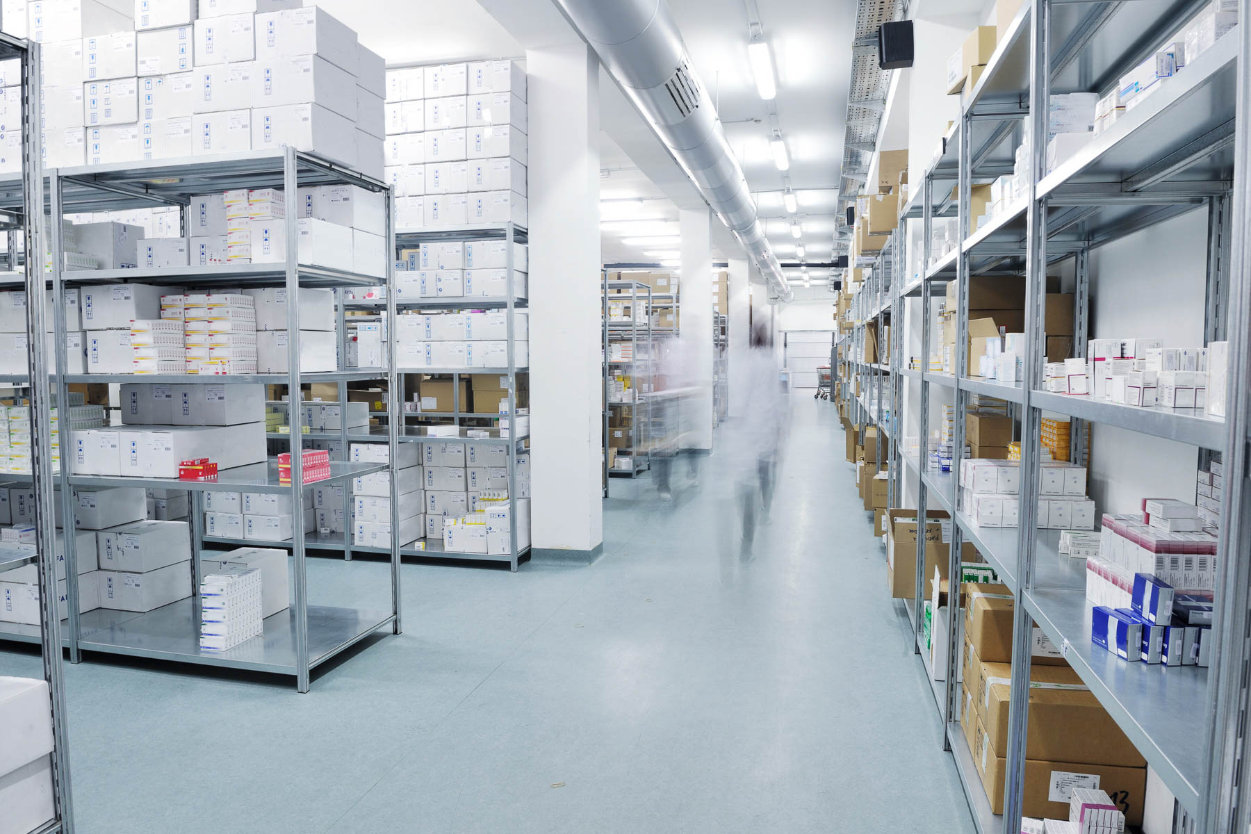 Rows of metal shelves in a warehouse, filled with boxes and packages, under fluorescent lighting.