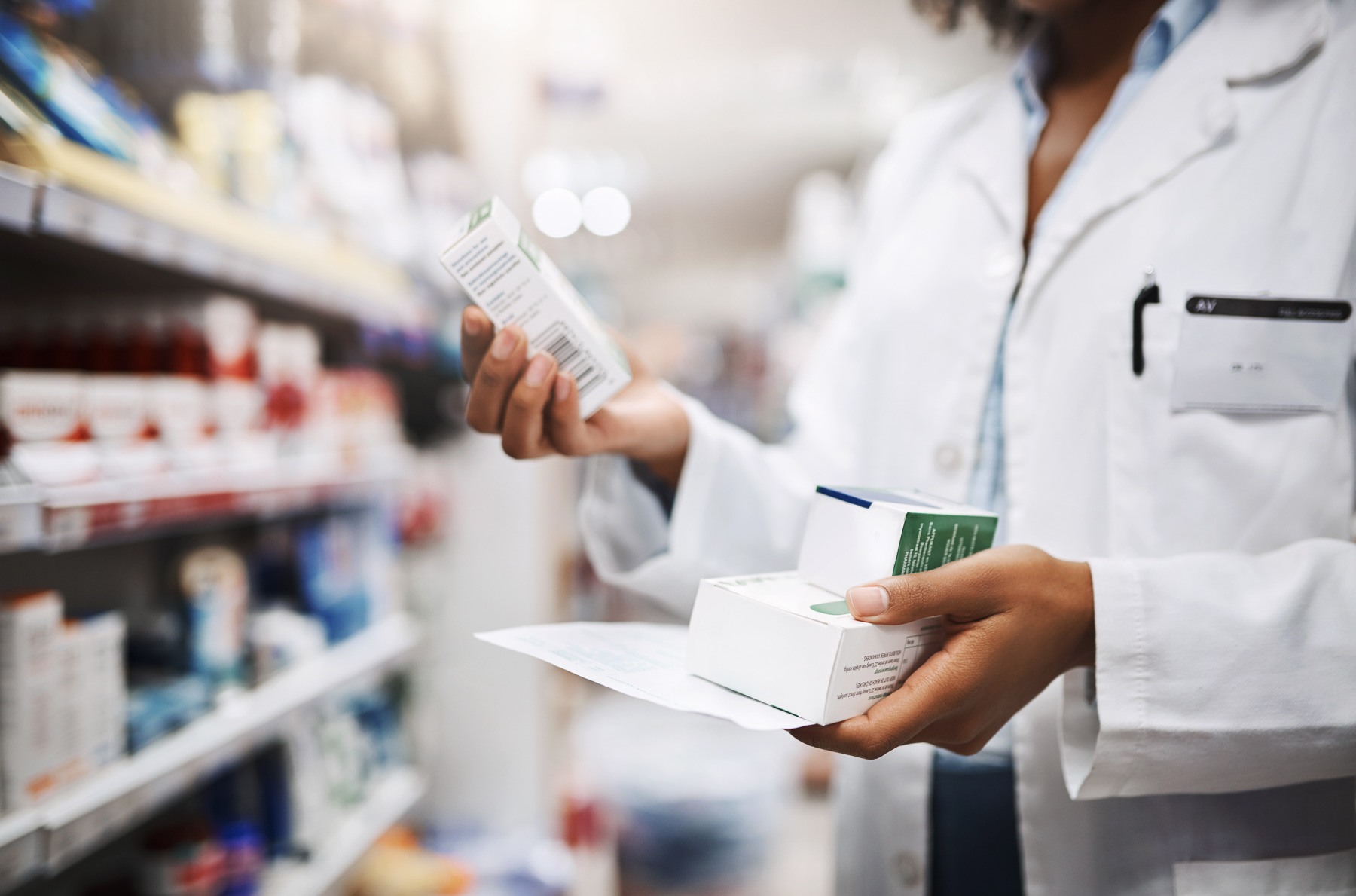 Pharmacist holding medication boxes and a prescription in a pharmacy aisle.