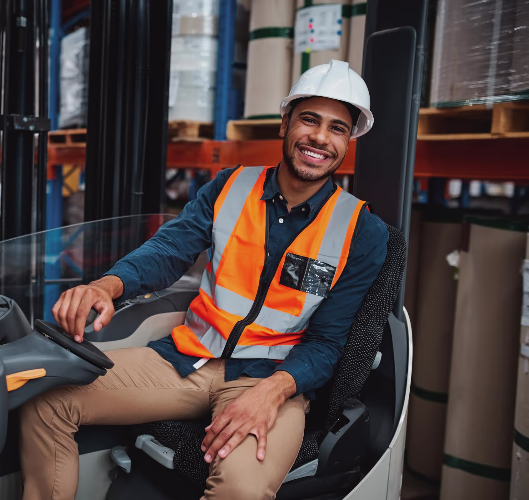 A man in a safety vest and hard hat sits smiling on a forklift in a warehouse.