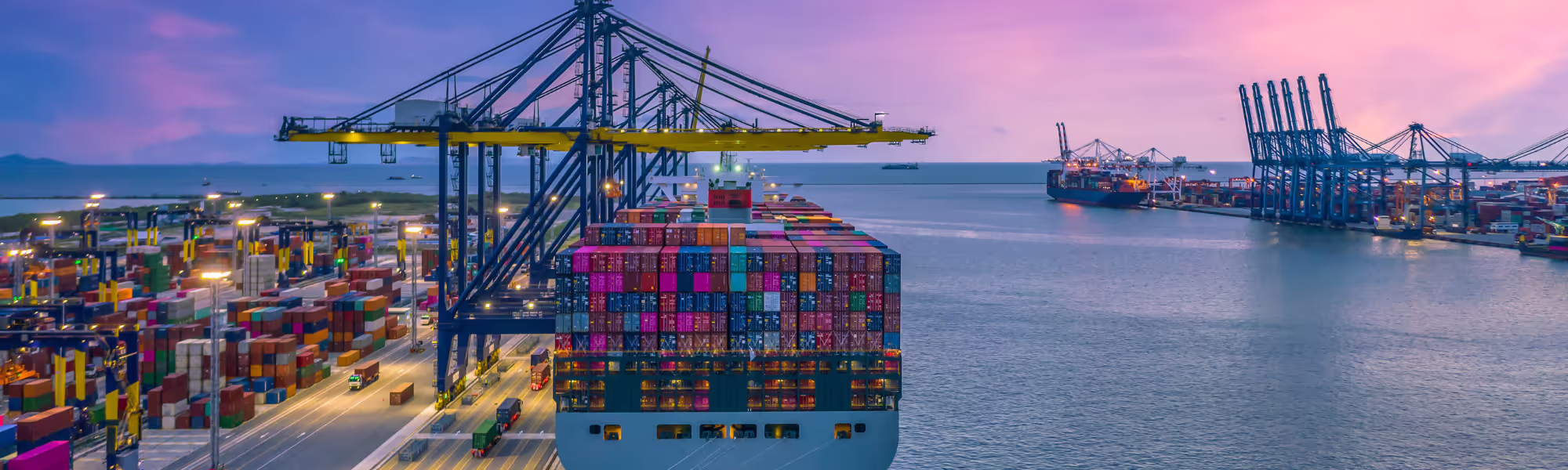 Cargo ship loaded with colorful containers docked at a busy port during sunset.