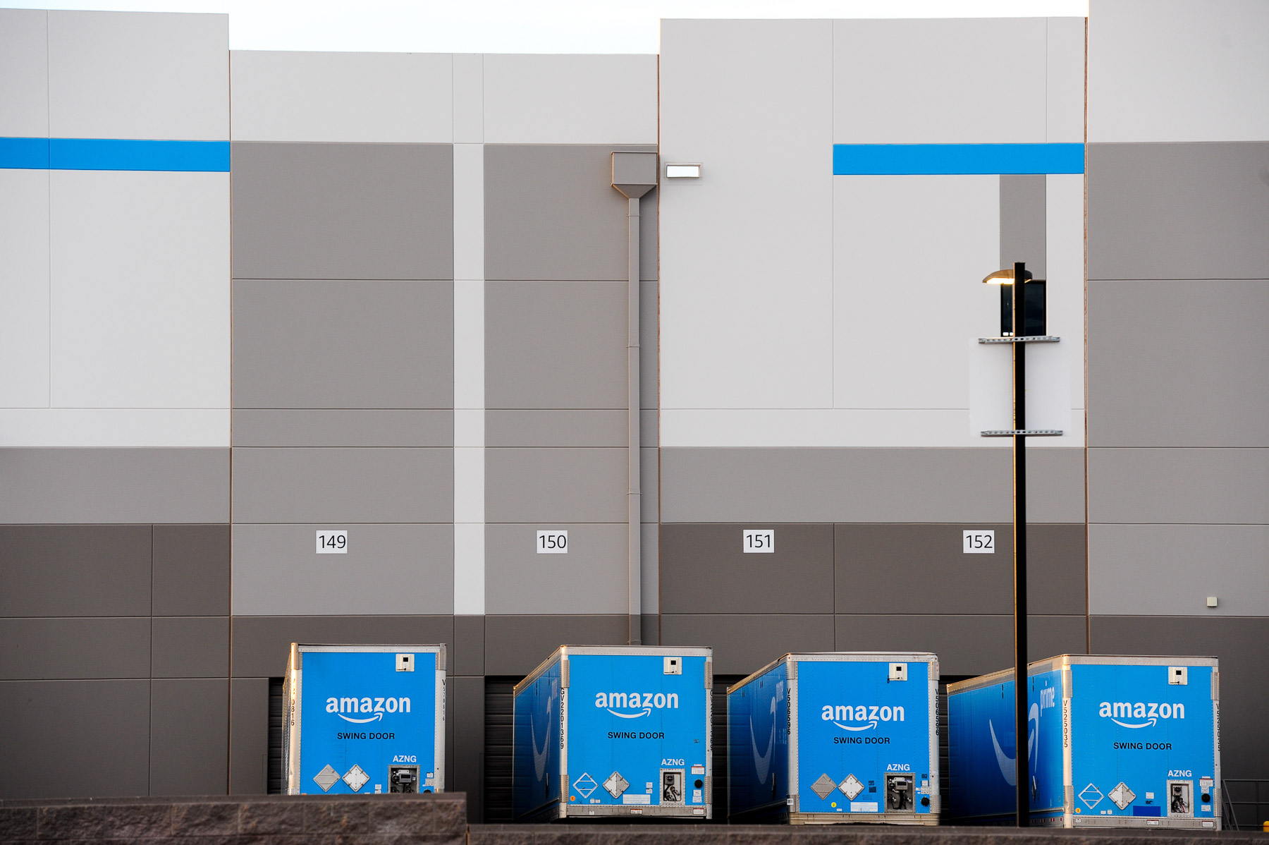 Four Amazon shipping containers are lined up outside a large gray and blue warehouse.