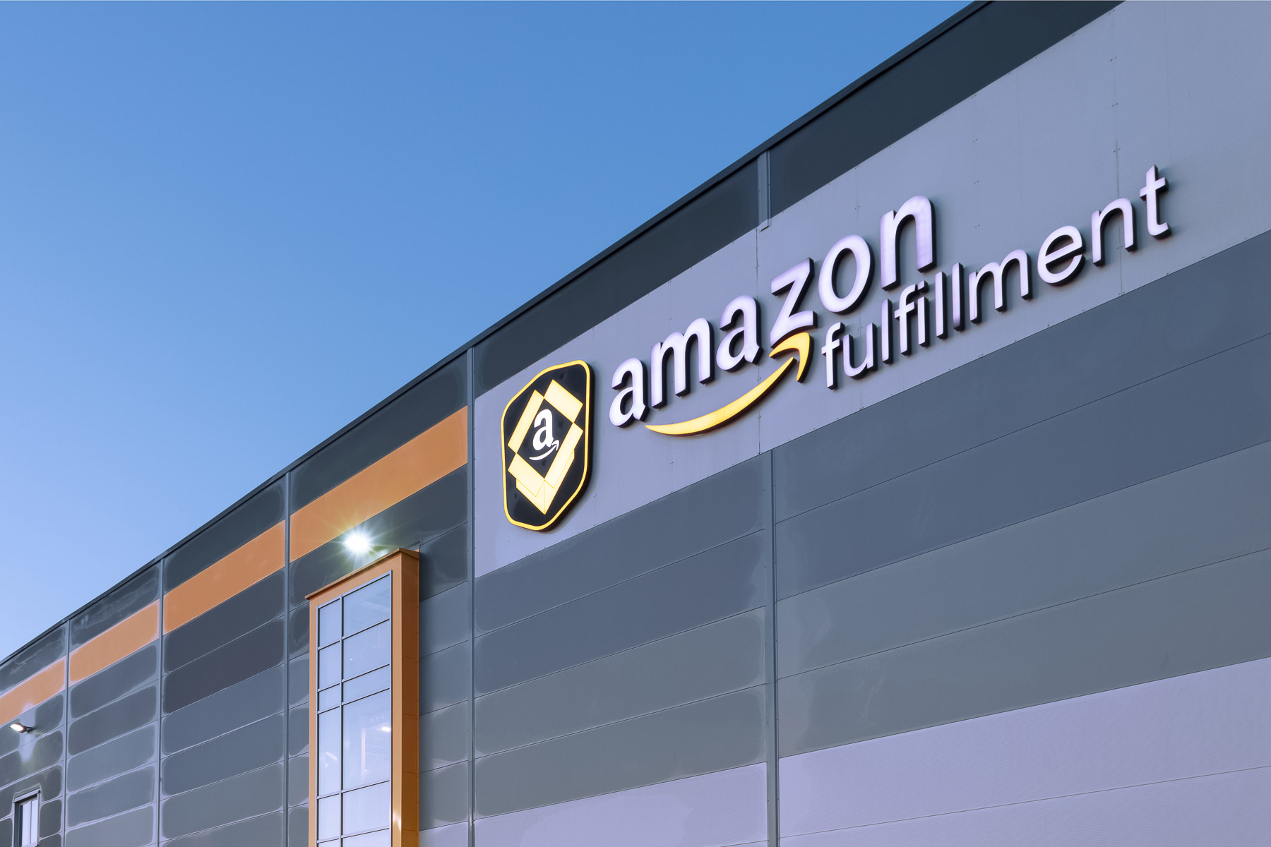 Sign on a building reads "Amazon Fulfillment." It's a gray industrial-style structure with a clear sky above.