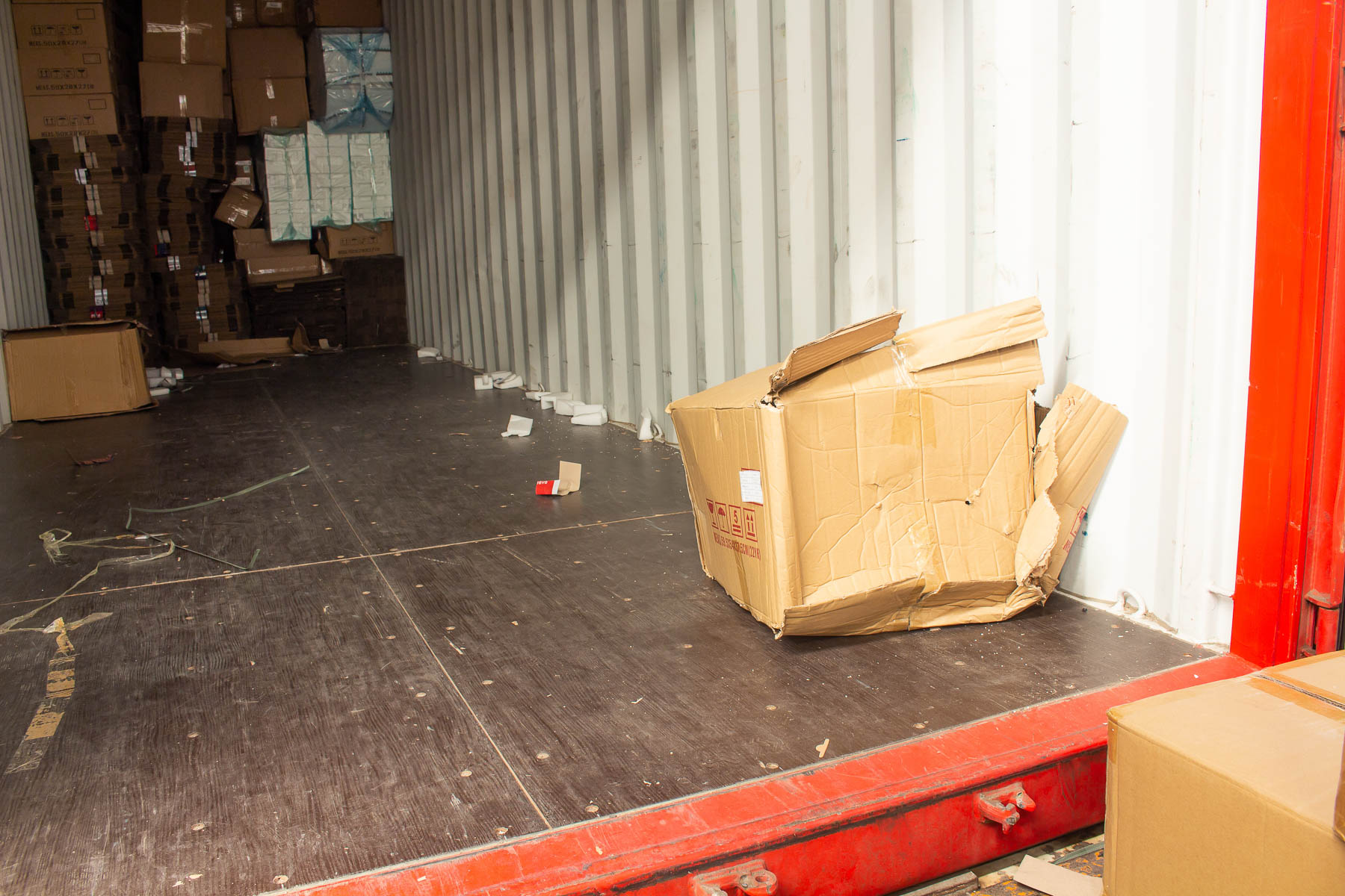 A damaged cardboard box sits at the edge of an open shipping container on a sunny day.
