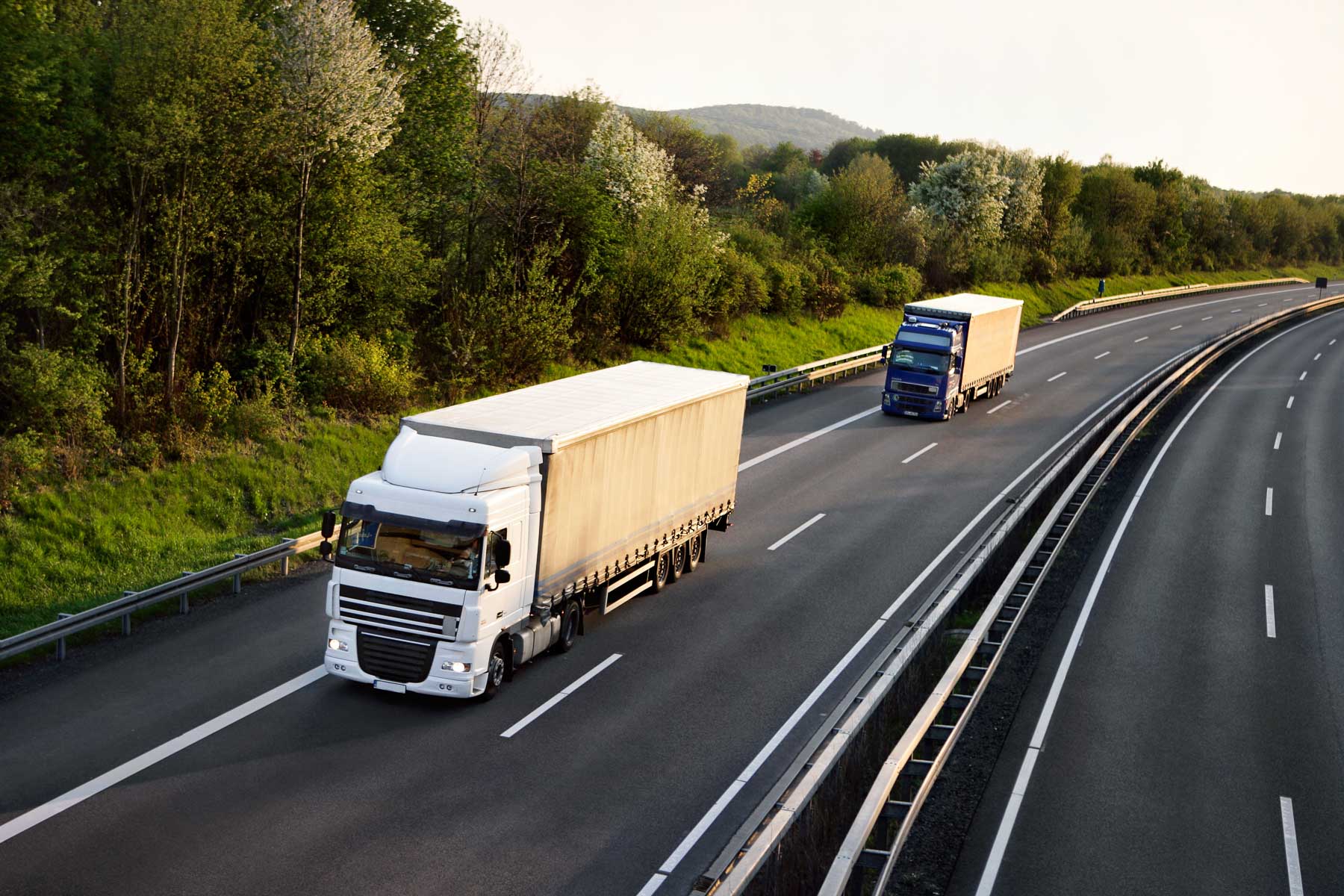 Two large trucks driving on a highway surrounded by trees and greenery.