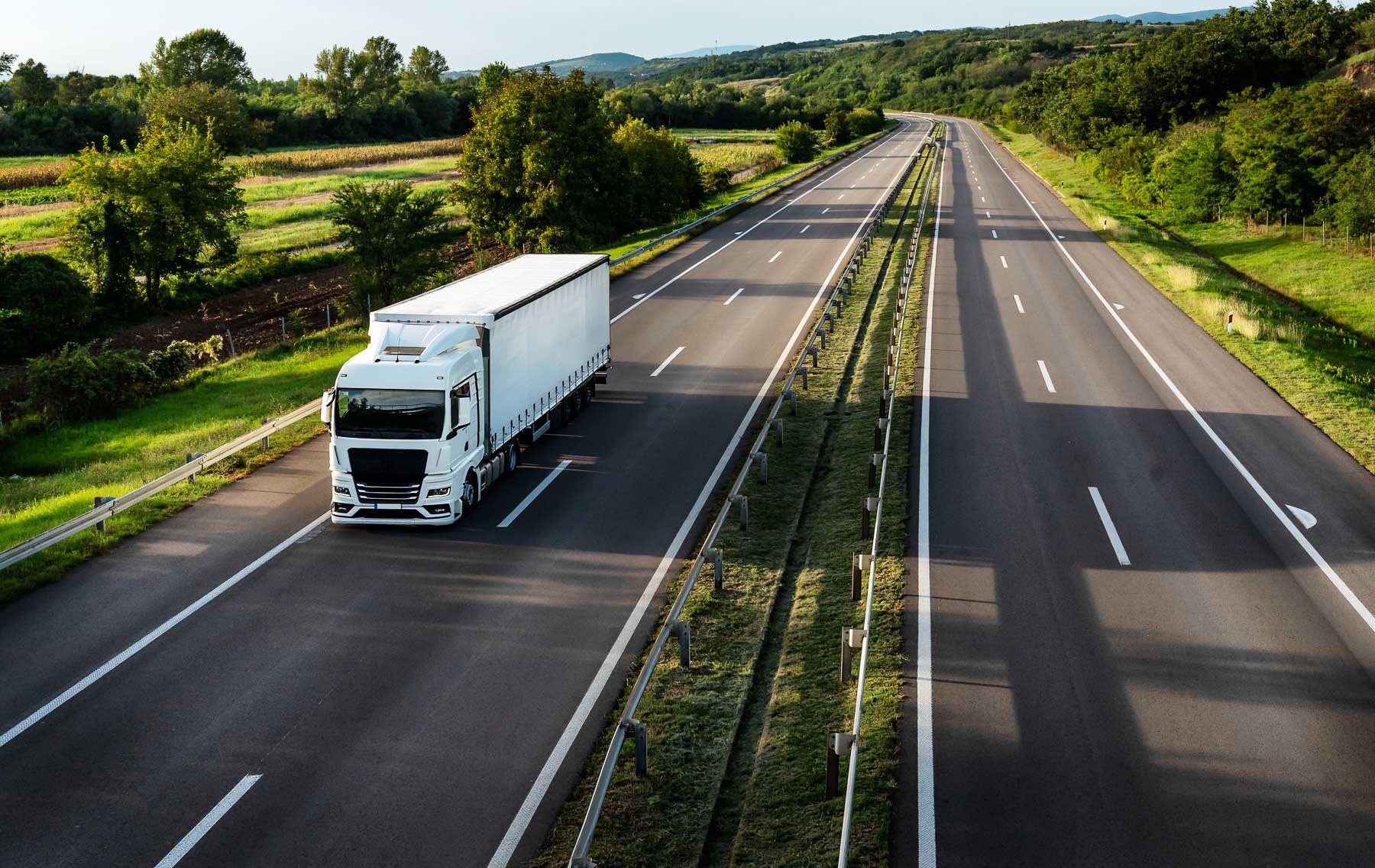 A white truck drives along an empty highway surrounded by green fields and trees.
