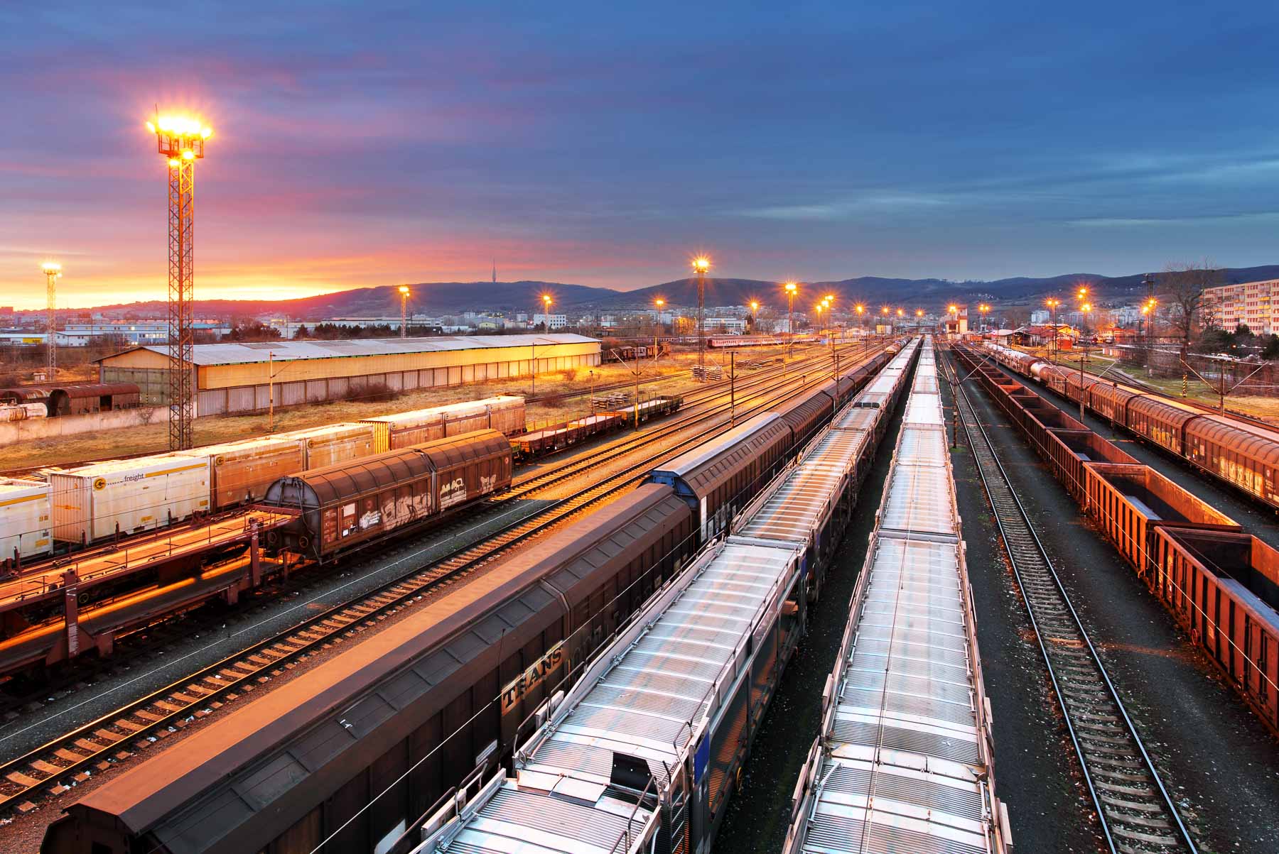 Train yard with multiple freight trains on tracks at sunset, under a cloudy sky.