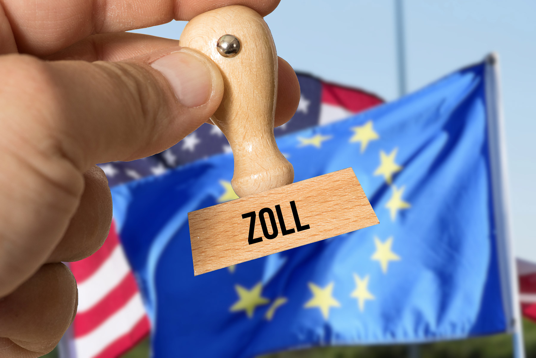 A hand holds a stamp marked "Zoll" in front of EU and US flags.