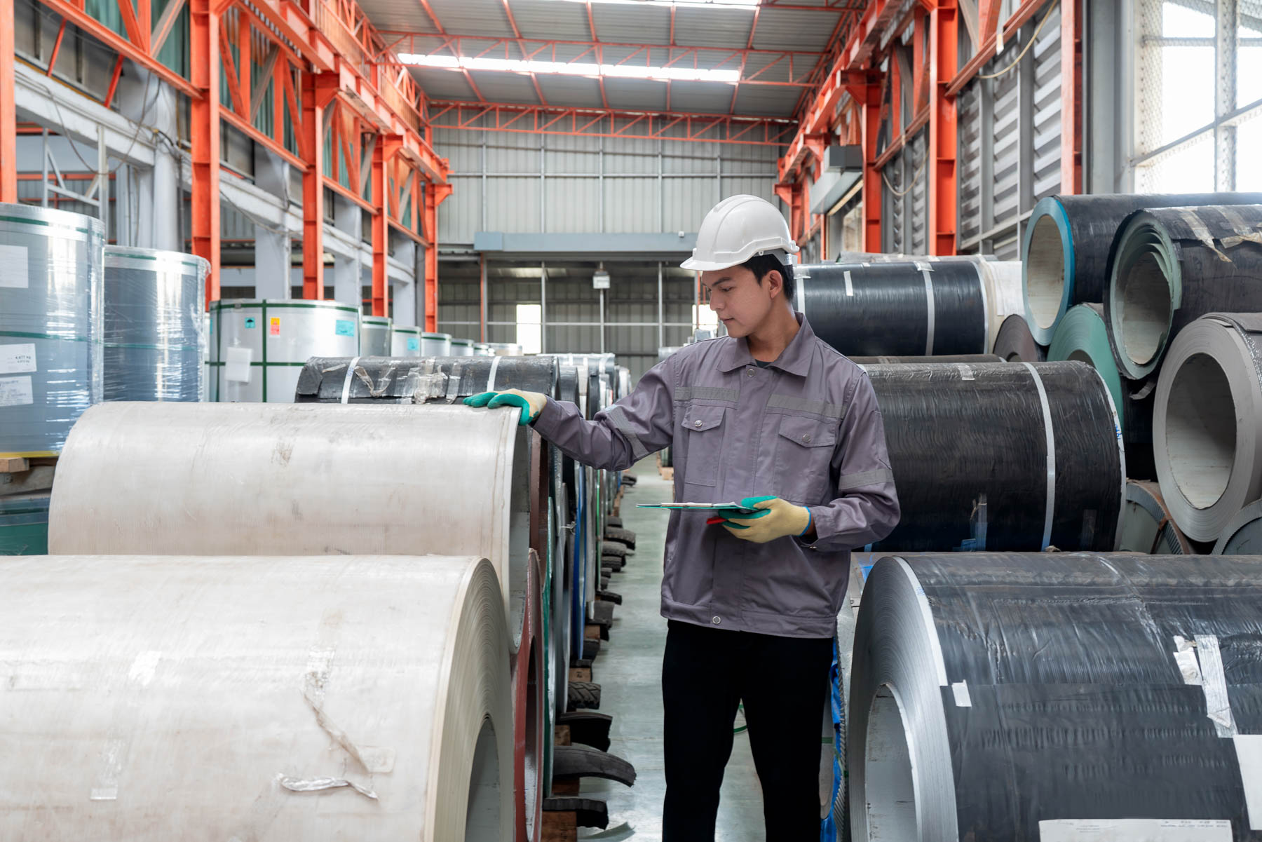 A worker with a hard hat inspects large metal coils in a warehouse.