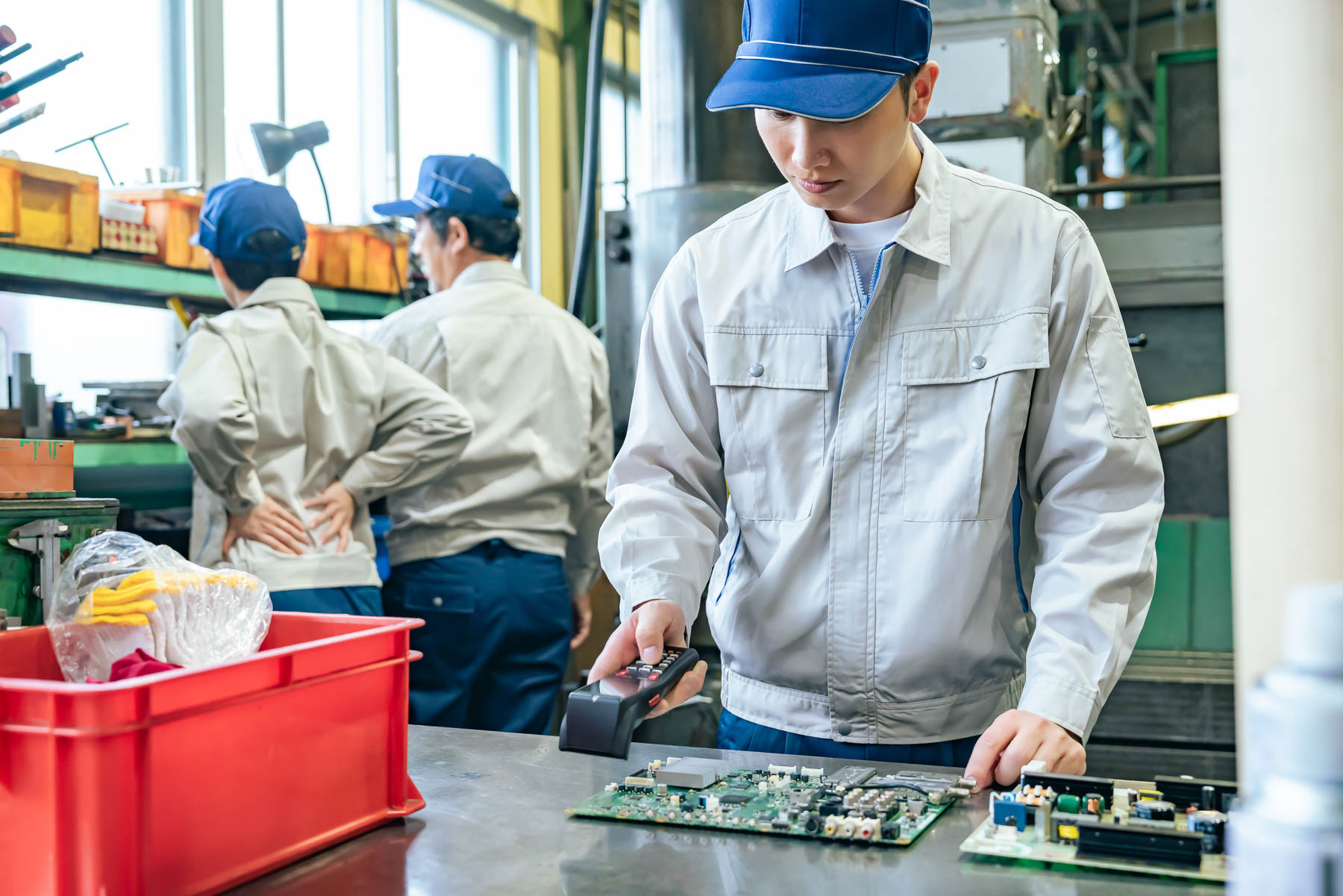 A worker in a uniform inspects a circuit board with a magnifying tool at a workstation.