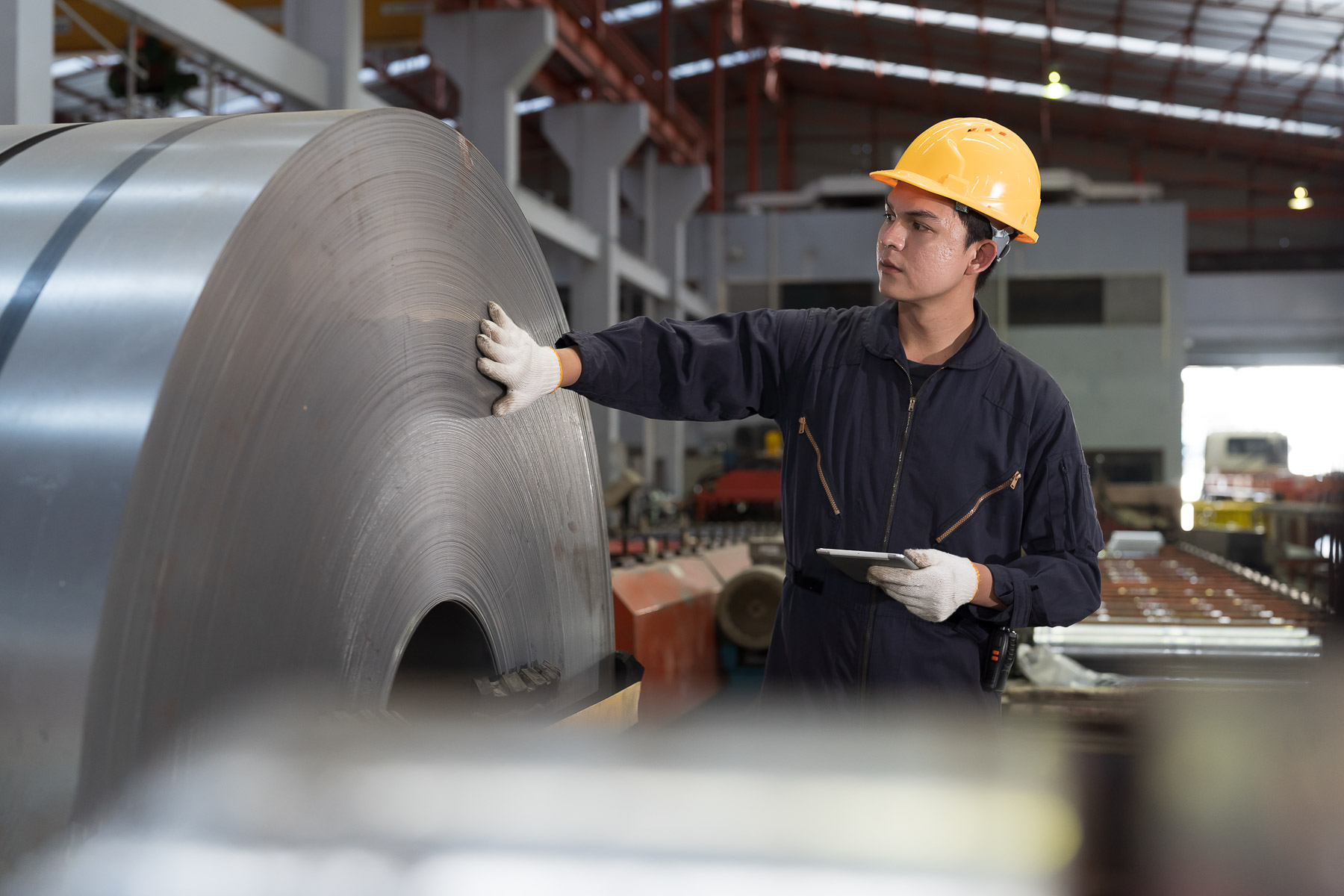 Worker in safety gear examines large metal coil in an industrial facility.