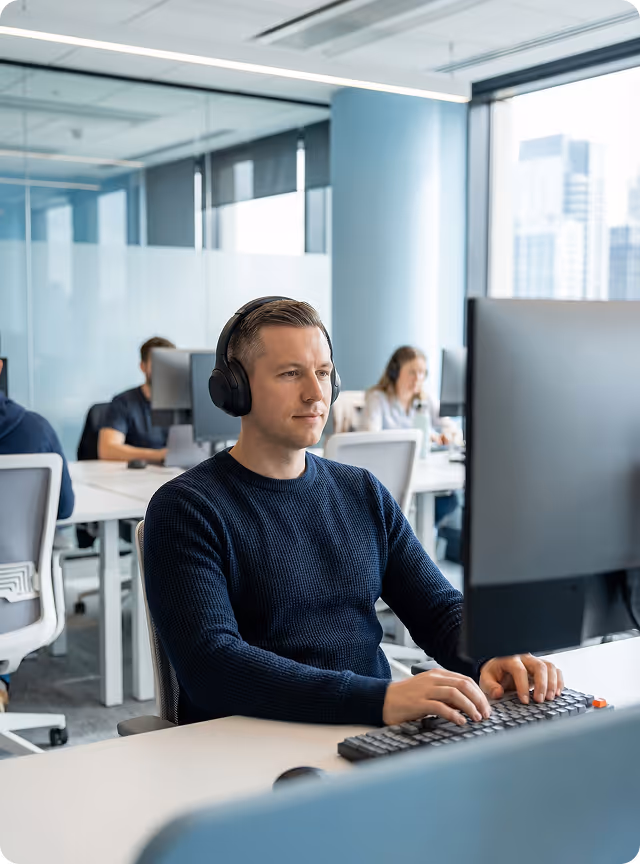 Man wearing headphones working on a computer keyboard in a modern office with colleagues in the background.