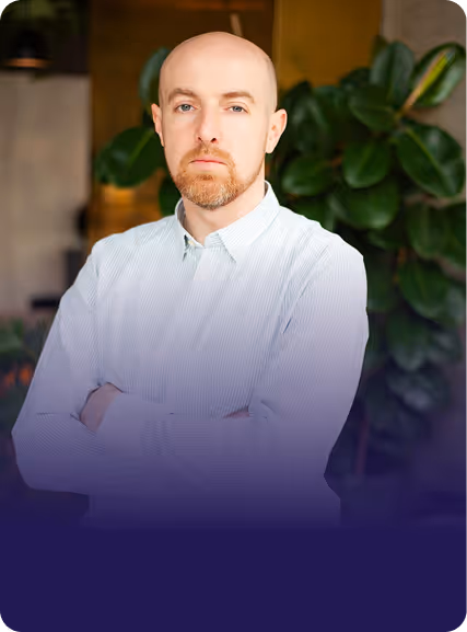 Portrait of a bald man with ginger beard wearing a light blue button-up shirt, standing with arms crossed indoors with green plants in the background.
