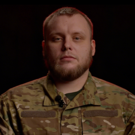 Чоловік у камуфляжній формі на темному фоні, дивиться в камеру з серйозним виразом обличчя