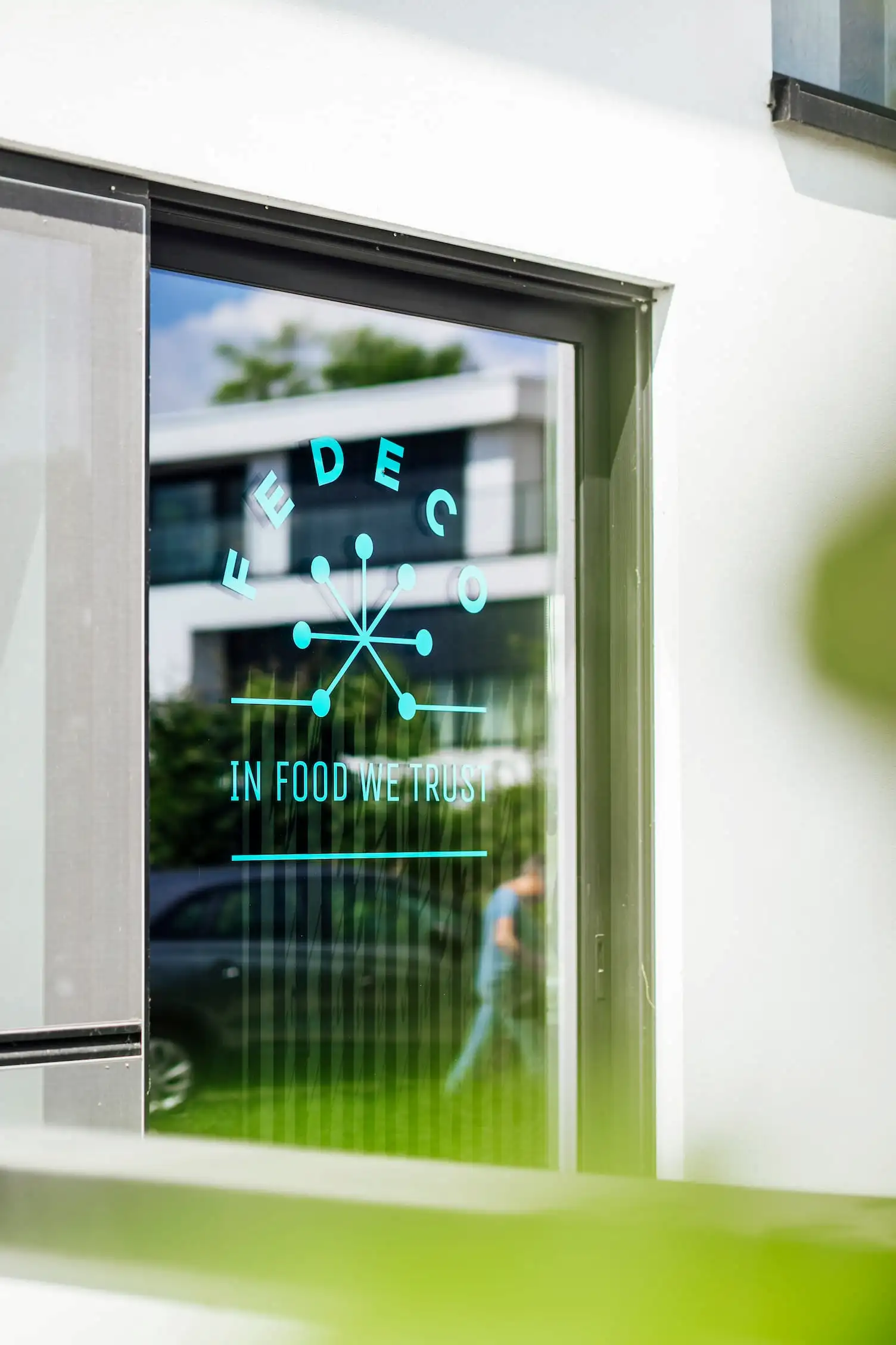 Glass door with blue logo reading 'FEDECO' and tagline 'IN FOOD WE TRUST' reflecting a modern building and a person walking outside.