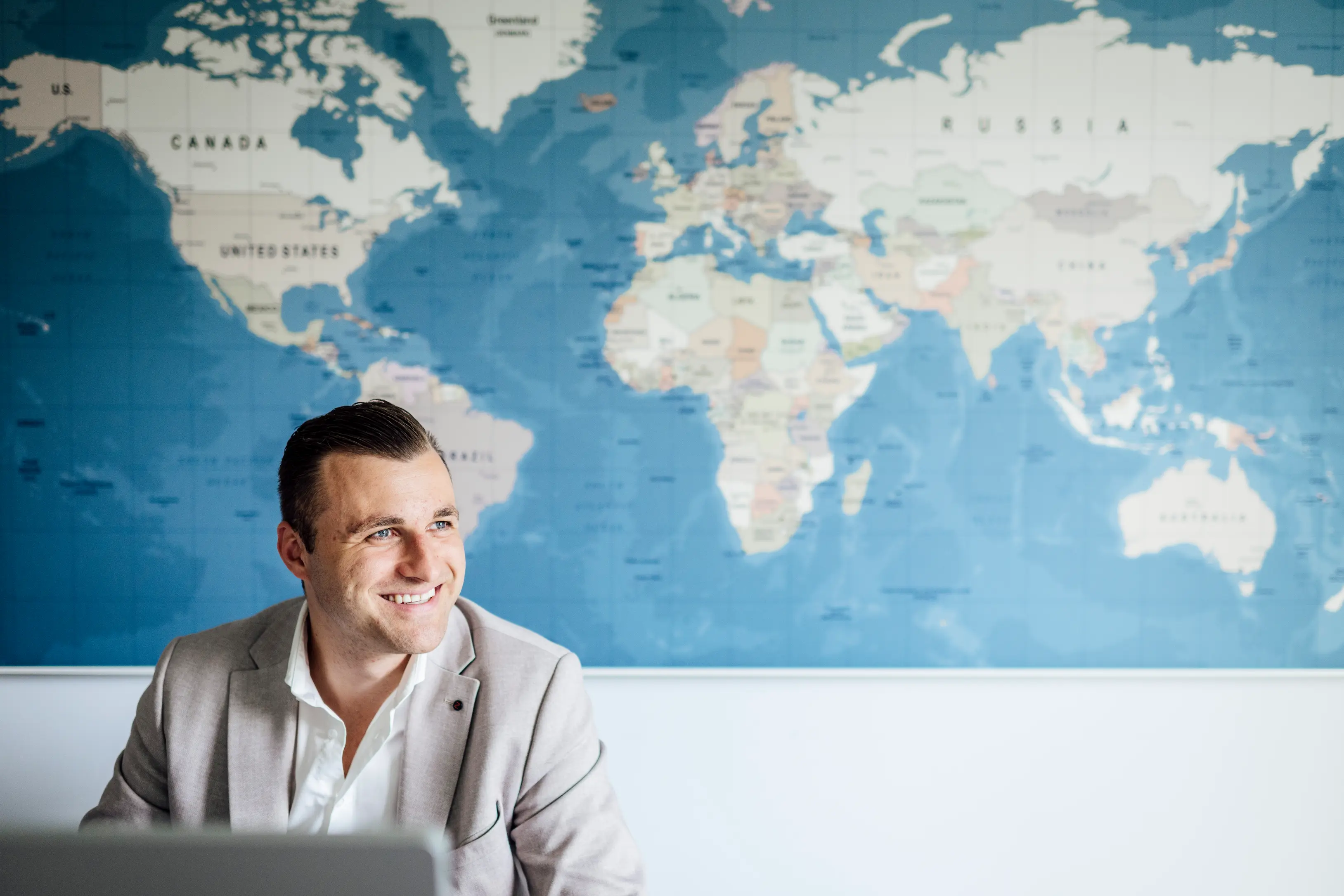 Smiling man in a light blazer sitting in front of a large world map on the wall.