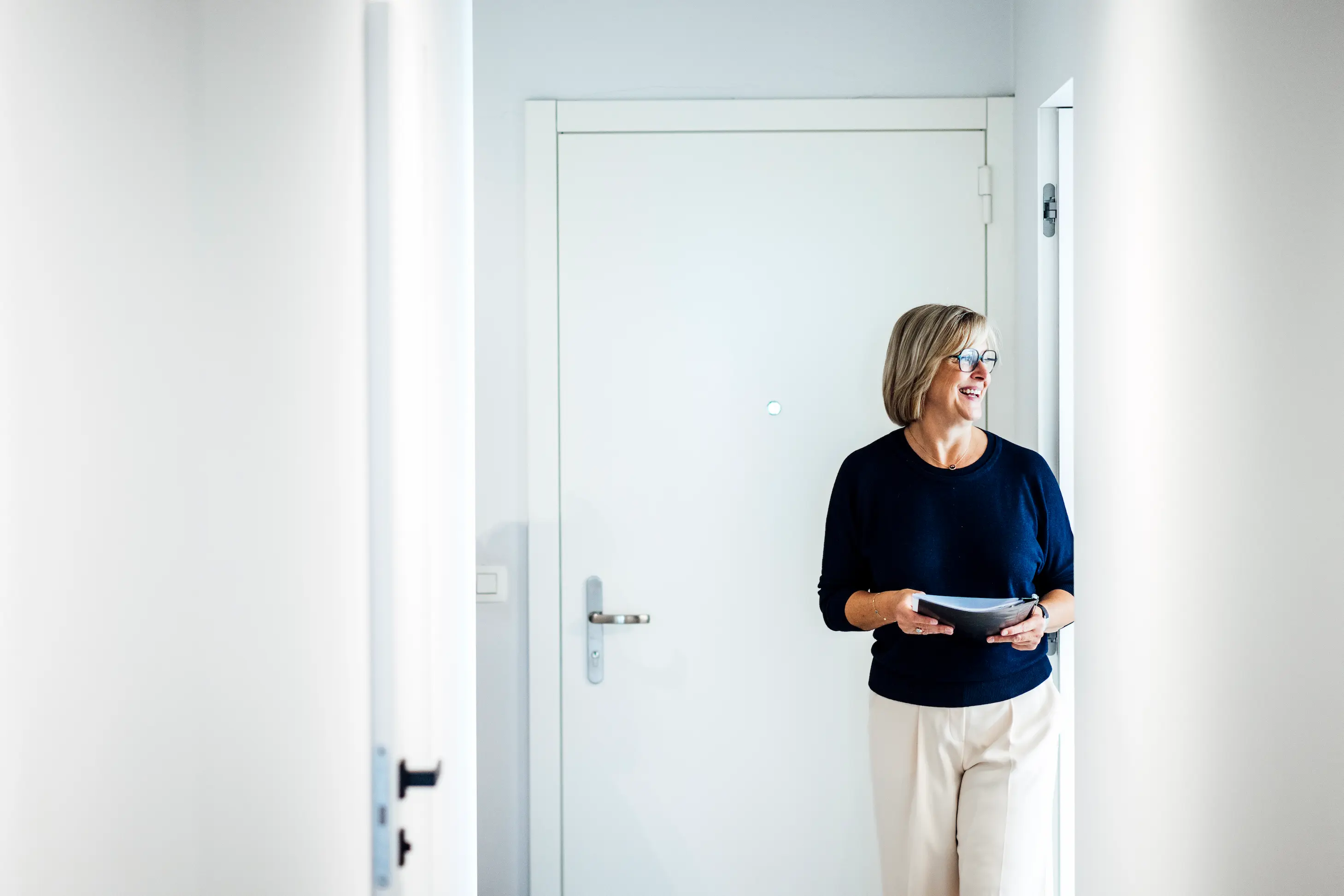 Smiling woman wearing glasses holds a folder while standing in a bright hallway near a closed white door.