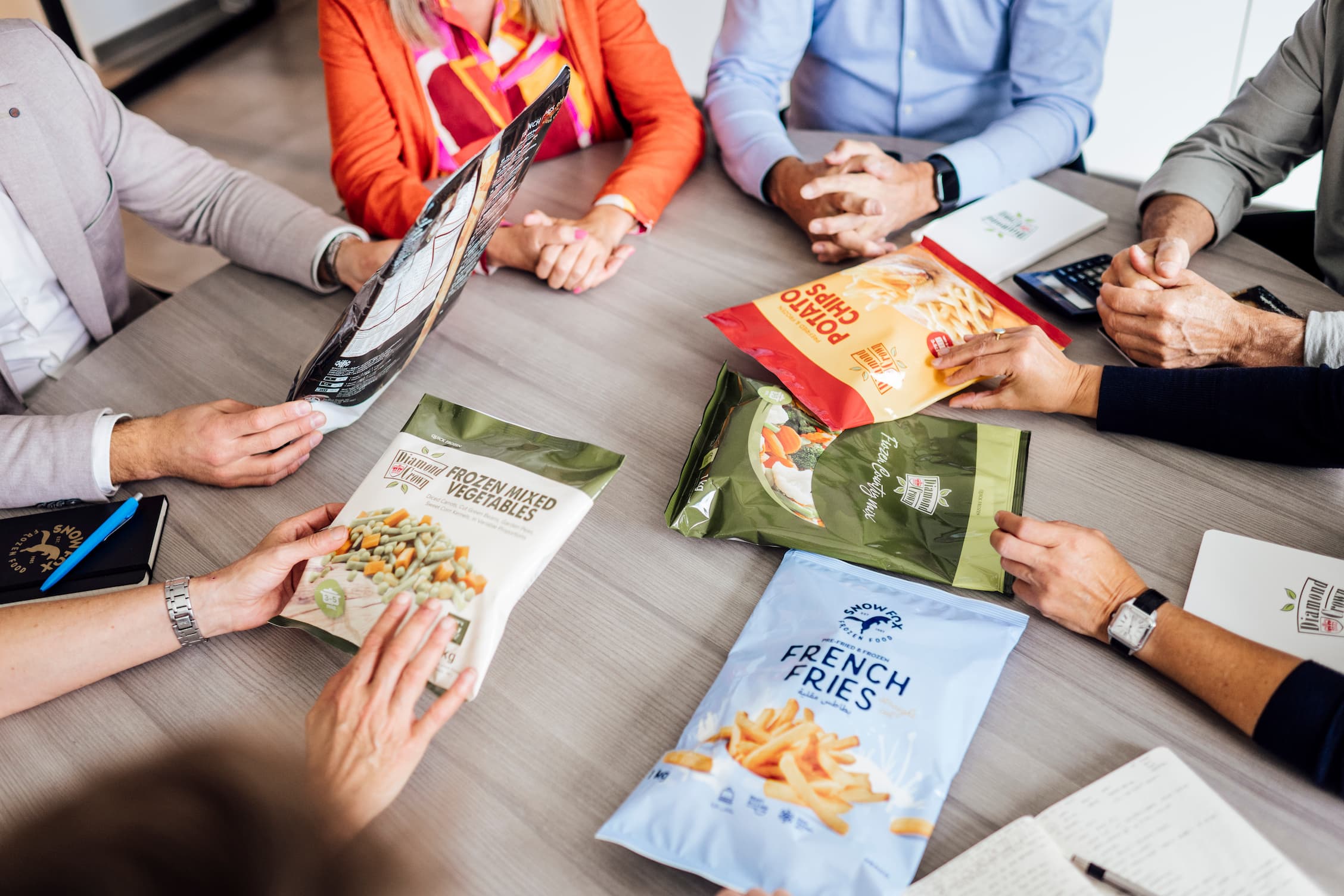 Group of people sitting around a table reviewing packaged frozen food products including mixed vegetables, french fries, and potato chips.