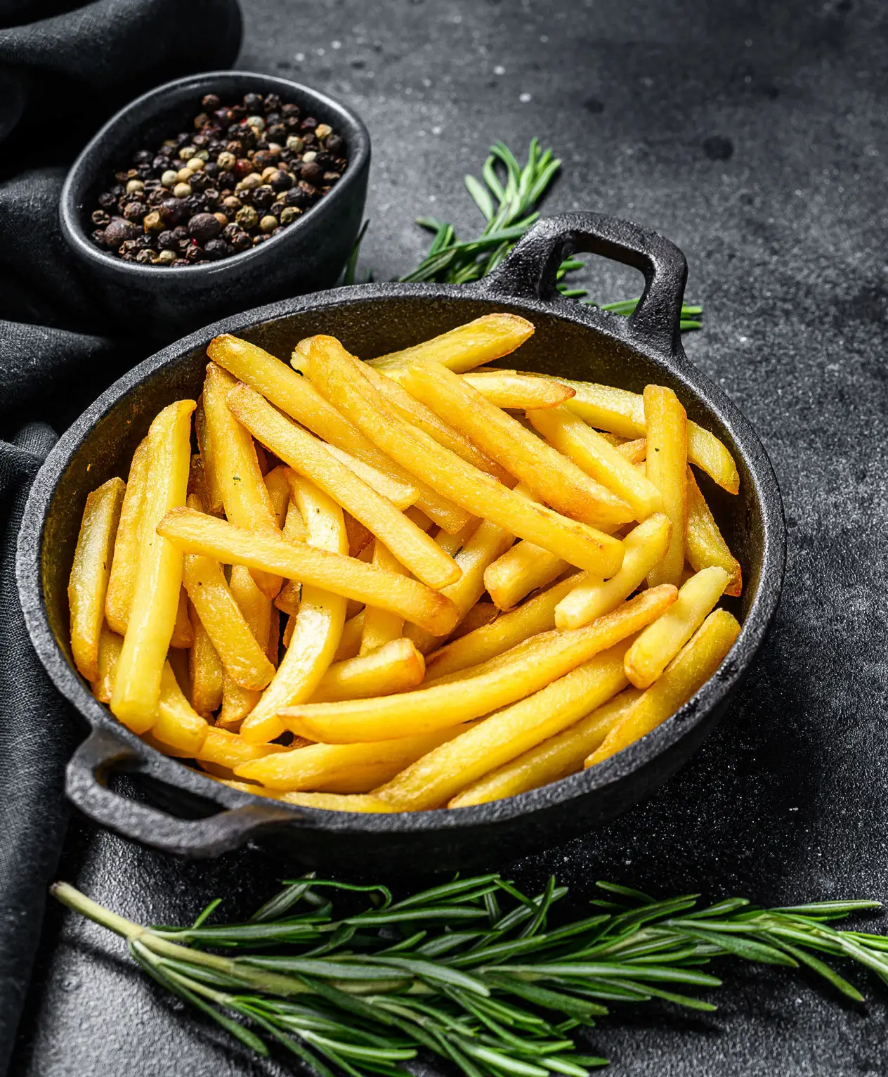 Cast iron pan filled with golden French fries, garnished with fresh rosemary and peppercorns nearby.