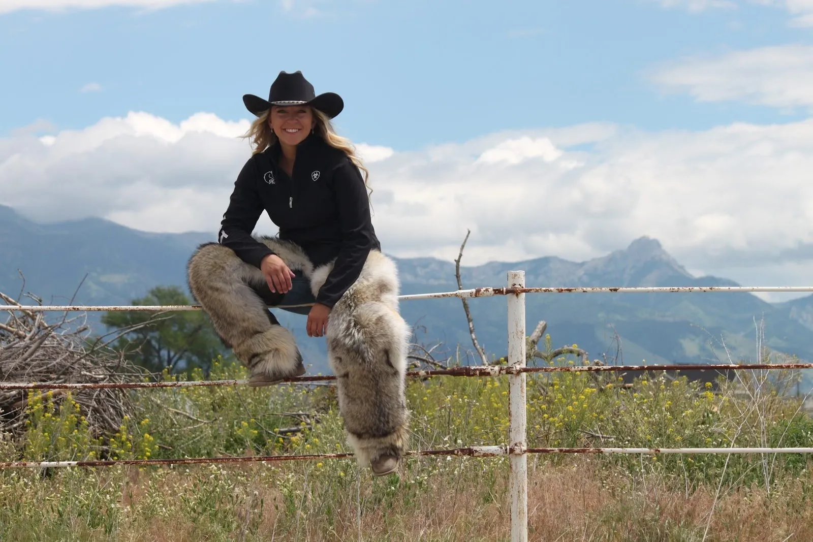 Woman sitting on a fence wearing a hat with mountains in the background.