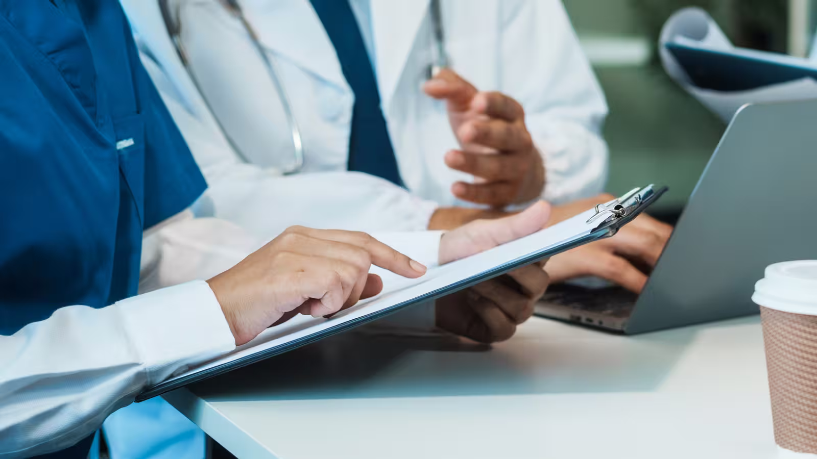 Clinicians reviewing a document on a clipboard at a meeting table, with a laptop open nearby.