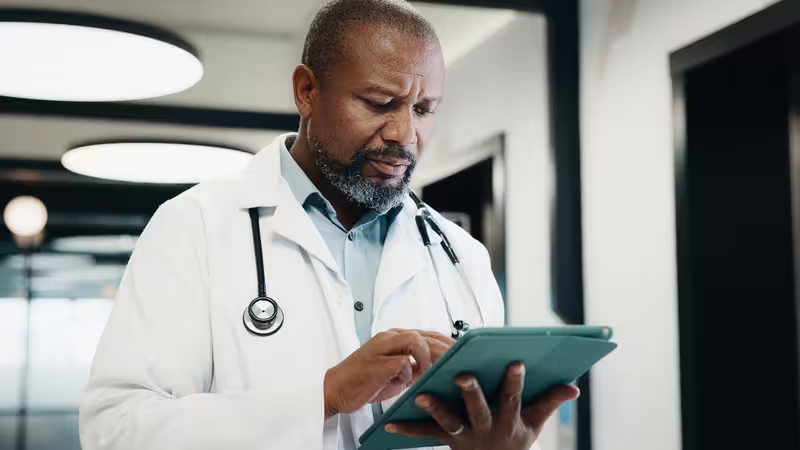 Doctor in a white coat and stethoscope using a tablet in a hospital corridor