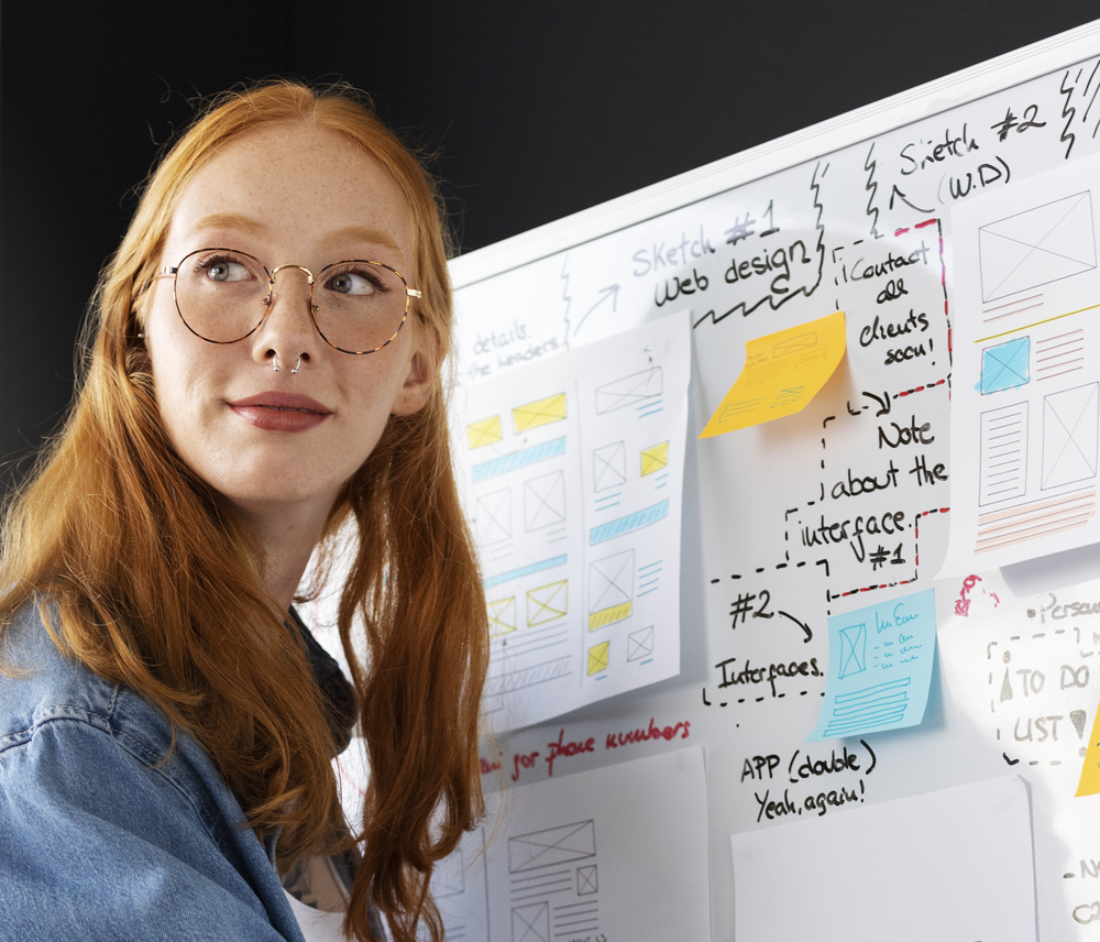 Woman with glasses beside a whiteboard covered in website wireframe sketches and sticky notes