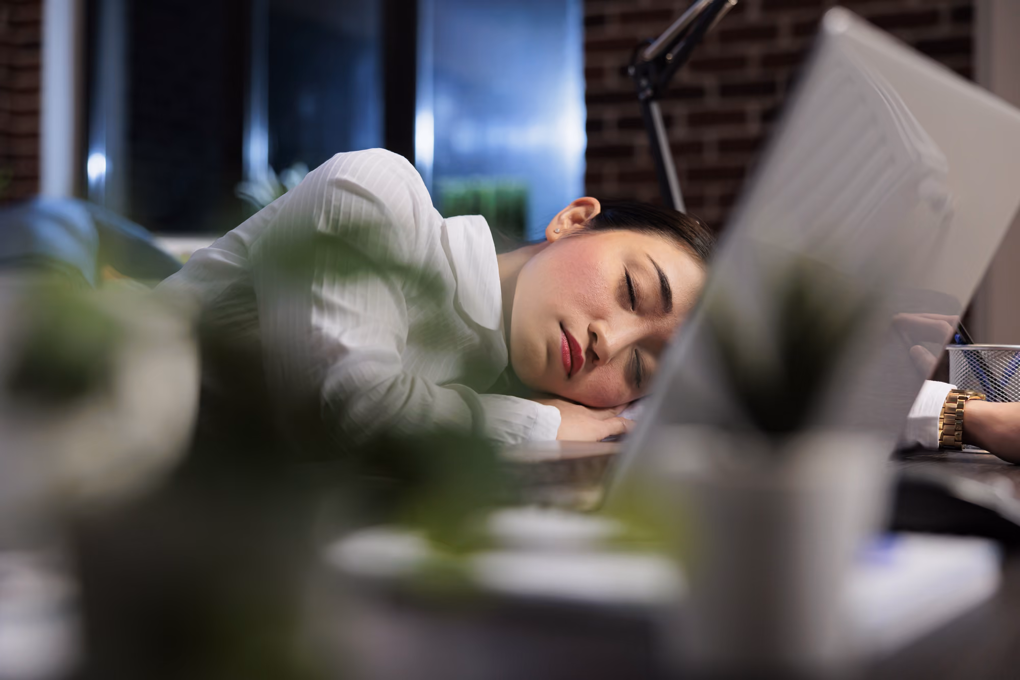 Tired woman asleep with her head resting on her arm at a desk beside a laptop