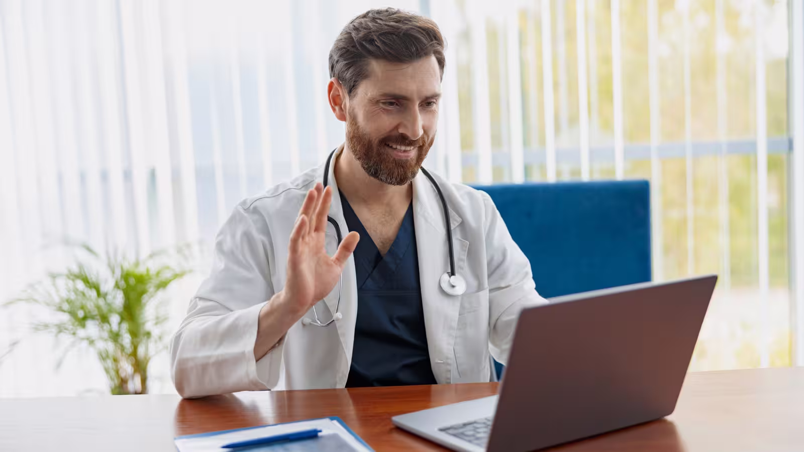 Smiling doctor in a white coat waving at a laptop during a video consultation.