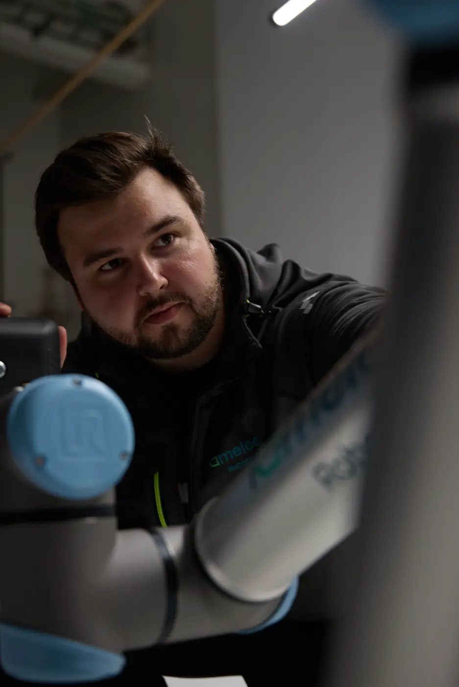 Mann med skjegg som undersøker en robotarm i et innendørs arbeidsmiljø.