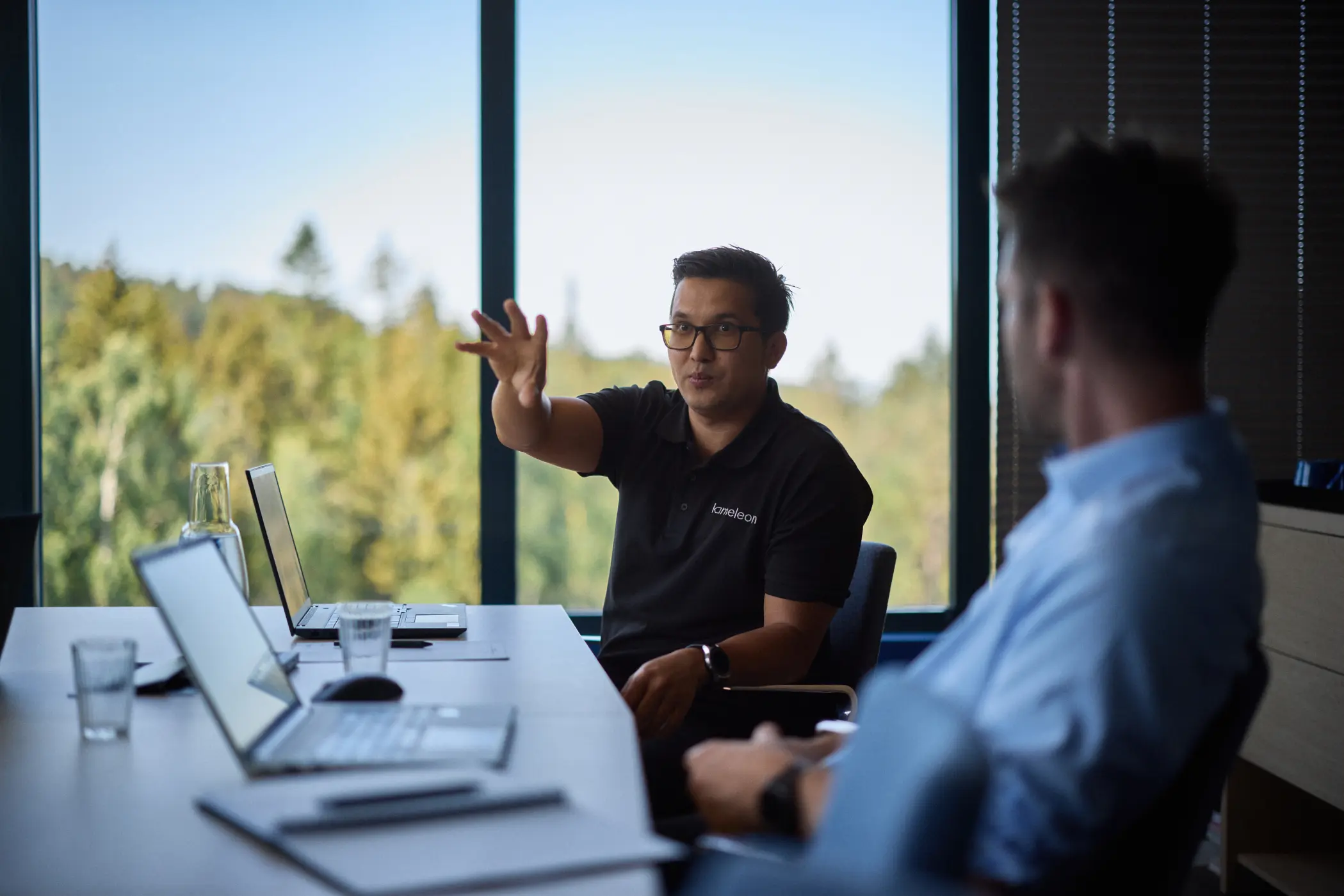 En mann i svart skjorte med briller gestikulerer mens han snakker med en annen mann i blå skjorte i et møterom med laptopper på bordet.