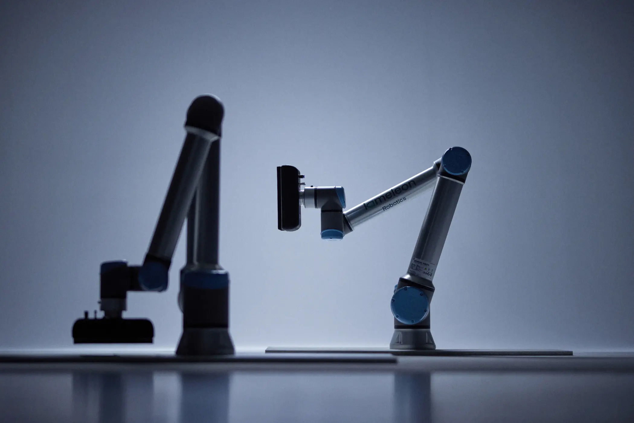 To industrielle robotarmer plassert på bord, med et minimalistisk og mørkt bakgrunnslys.
