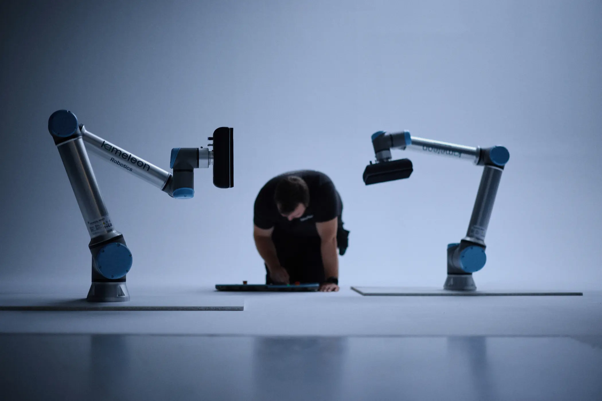 En person kneler på gulvet mellom to industrirobotar med blåarmer i eit minimalistisk, blått opplyst rom.