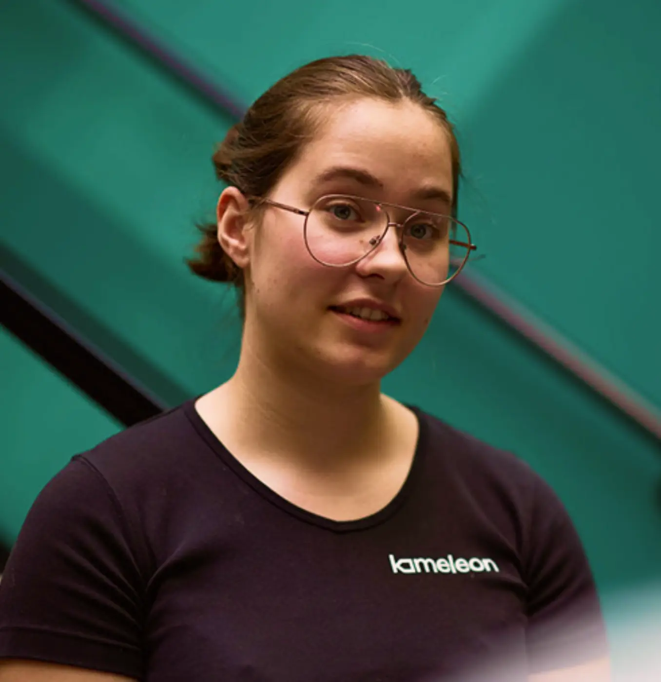 Kvinne med briller og mørk t-skjorte med teksten 'kameleon' foran en grønn bakgrunn.
