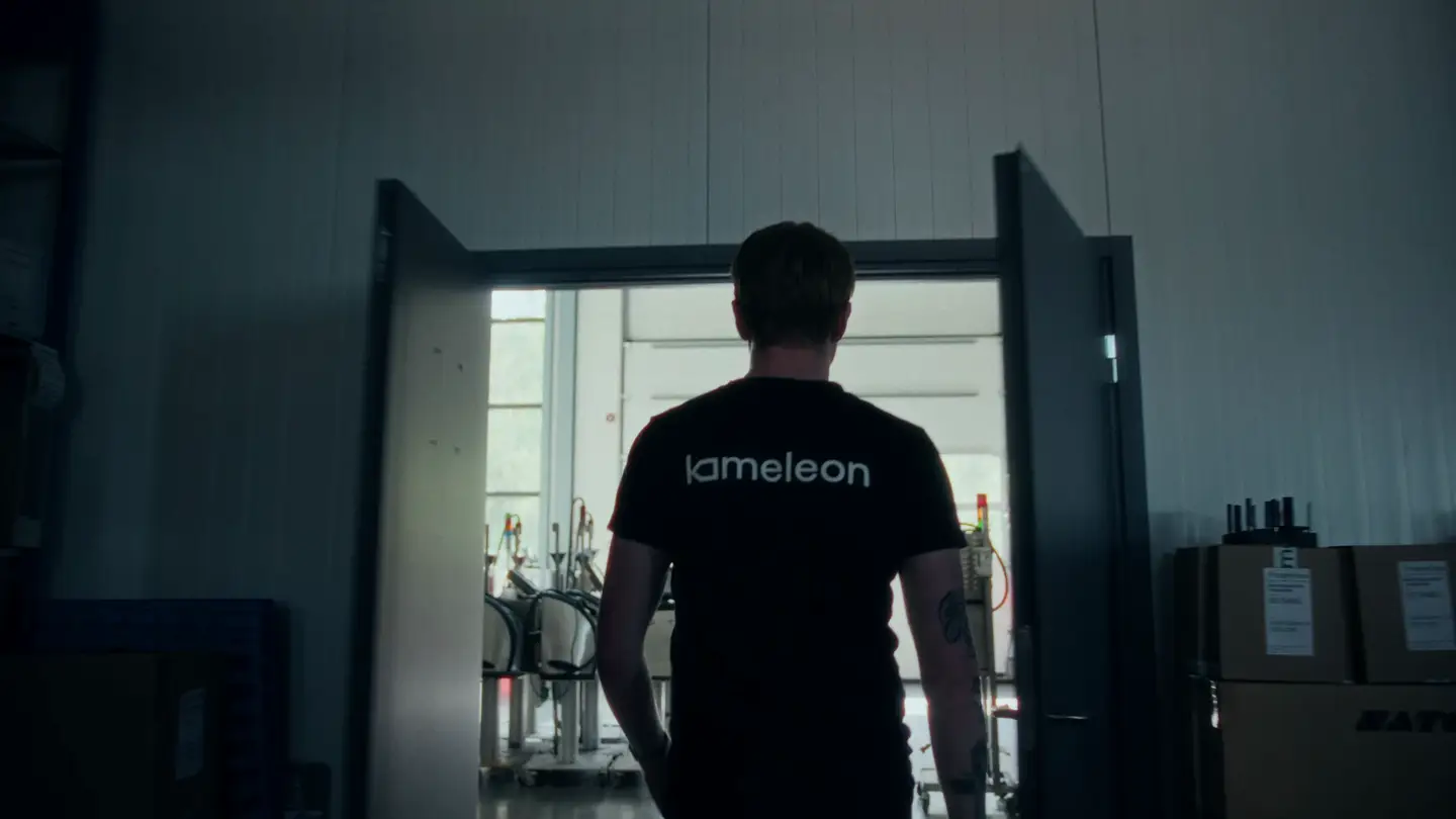 Person med svart t-skjorte med teksten 'kameleon' sett bakfra går gjennom en åpen dør inn i et lyst rom med teknisk utstyr.