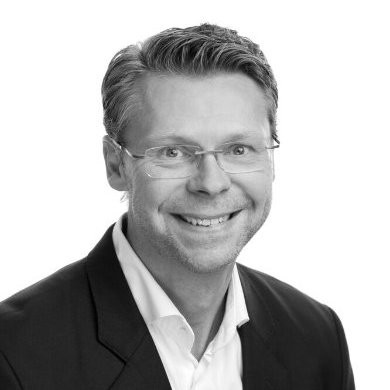 Portrett av en smilende mann med briller, mørk dressjakke og hvit skjorte mot hvit bakgrunn.