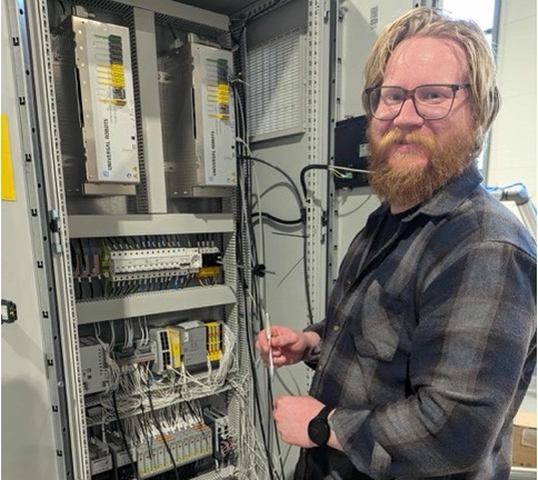 Smilende mann med briller og skjegg som står foran et åpent elektrisk kontrollpanel med mange ledninger.