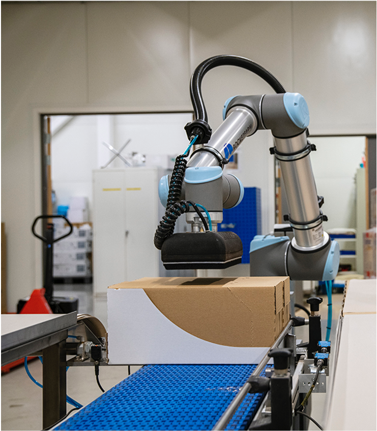 Industriell robotarm som løfter en pappeske på et bånd i en fabrikk.