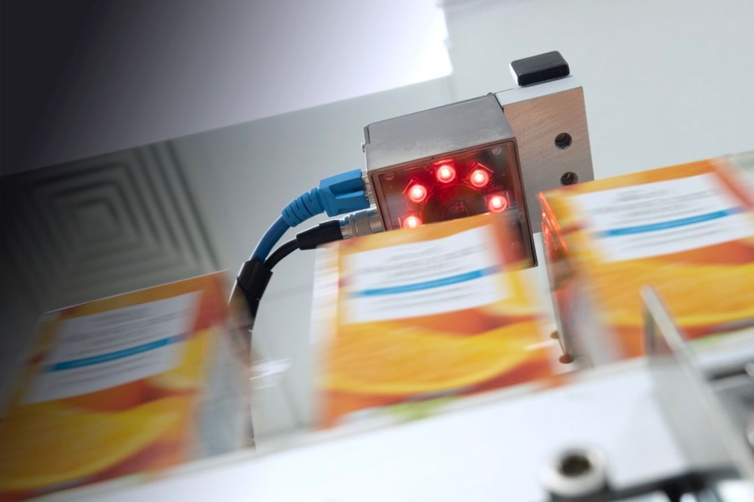 Industriell kamerasensor med røde lys som skanner bevegelige oransje emballasjer på et samlebånd.